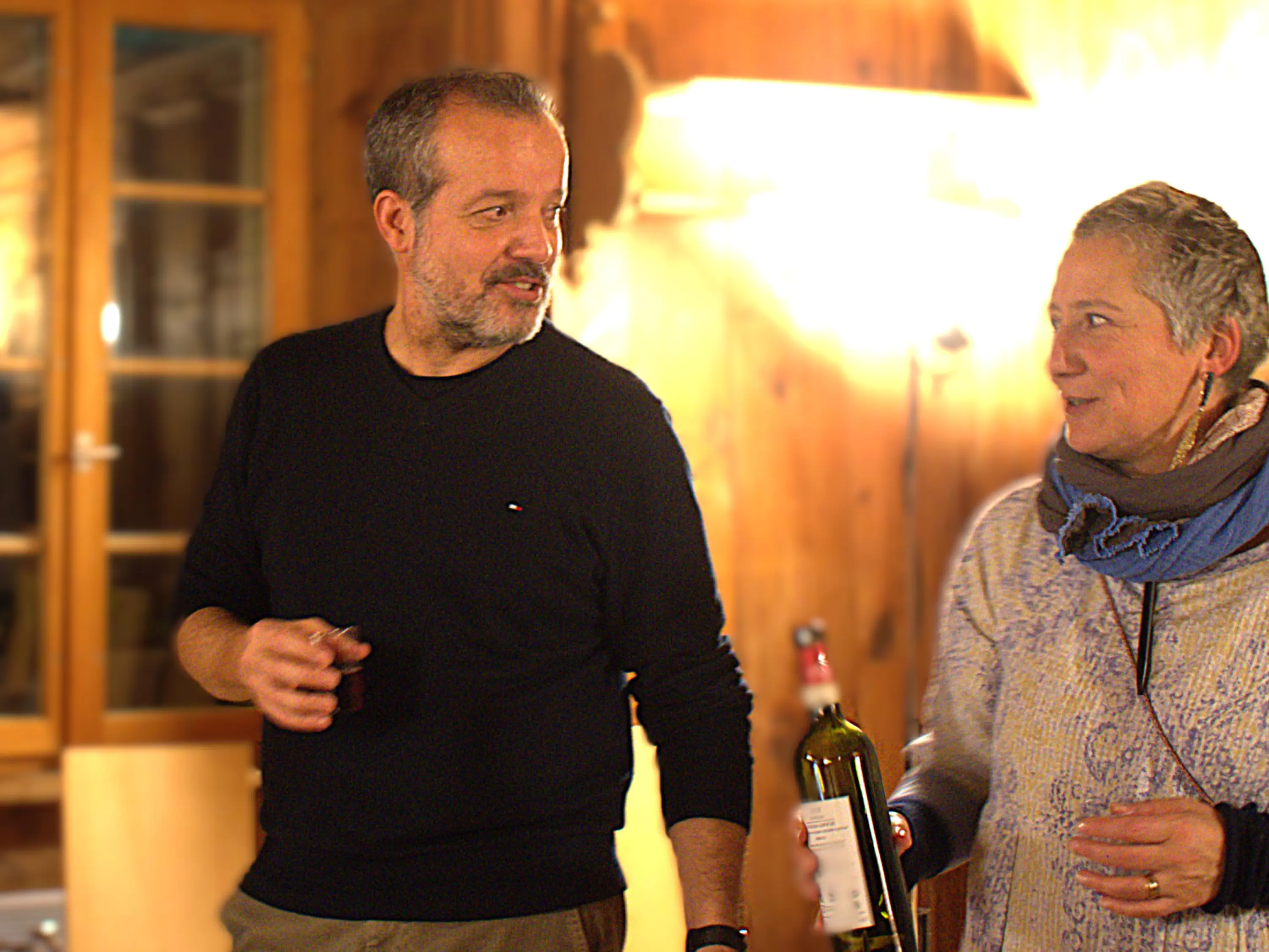 Due persone in una stanza accogliente, una con un bicchiere in mano e l'altra che mostra una bottiglia di vino. Sullo sfondo si vedono pareti in legno e luci calde.