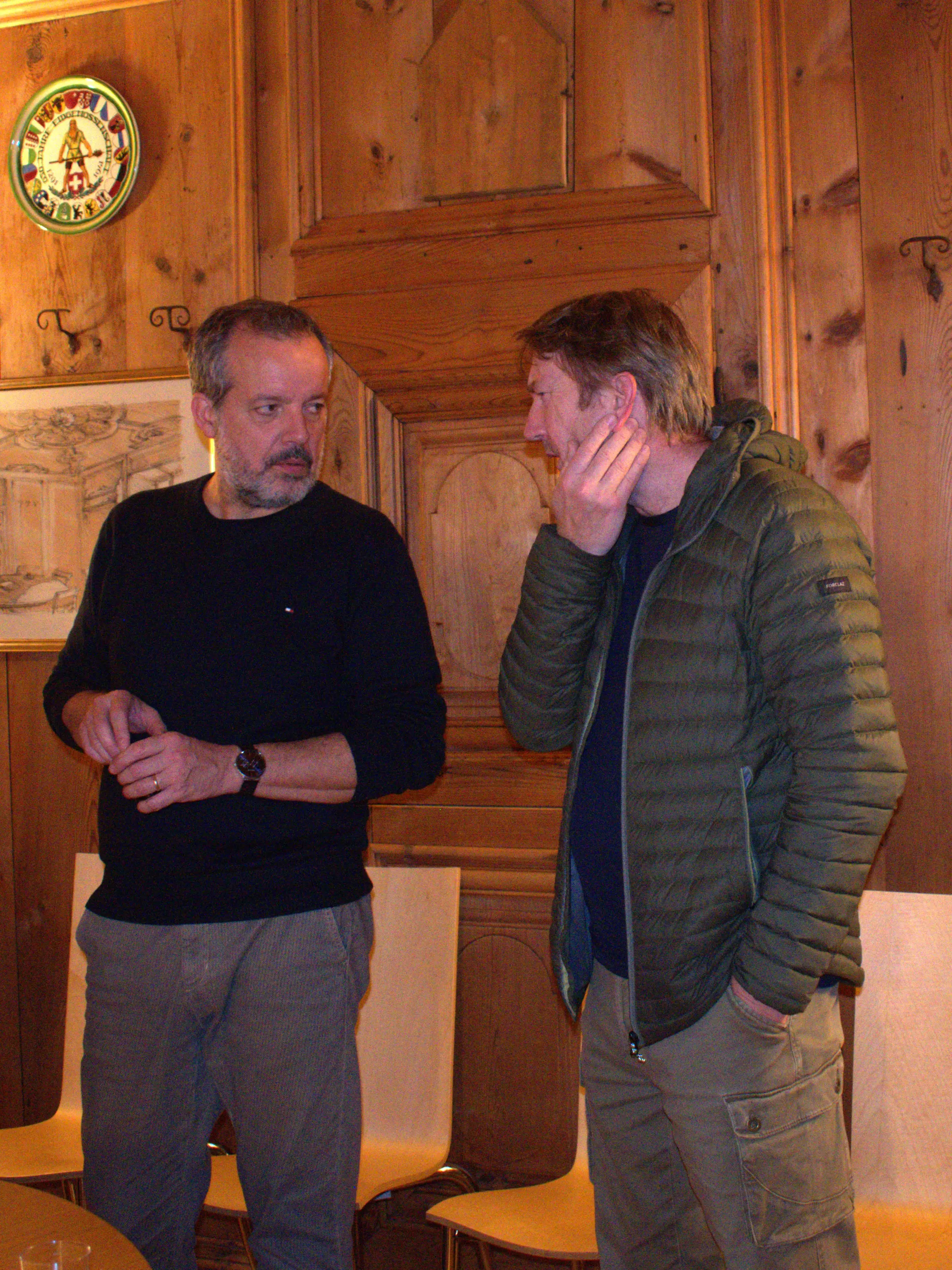 Due uomini in conversazione in una stanza con pareti in legno. Uno indossa un maglione scuro e l'altro una giacca verde. Entrambi sembrano pensierosi mentre discutono.