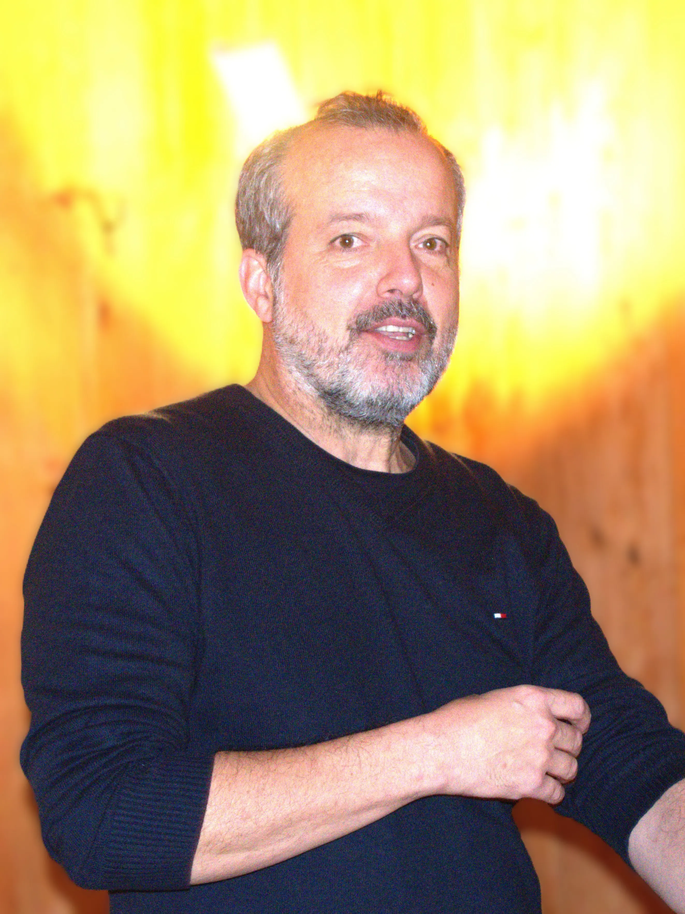 Un uomo con barba grigia e capelli castani, indossa un maglione nero. Sta parlando con un sorriso, mentre si trova di fronte a uno sfondo luminoso e sfocato.