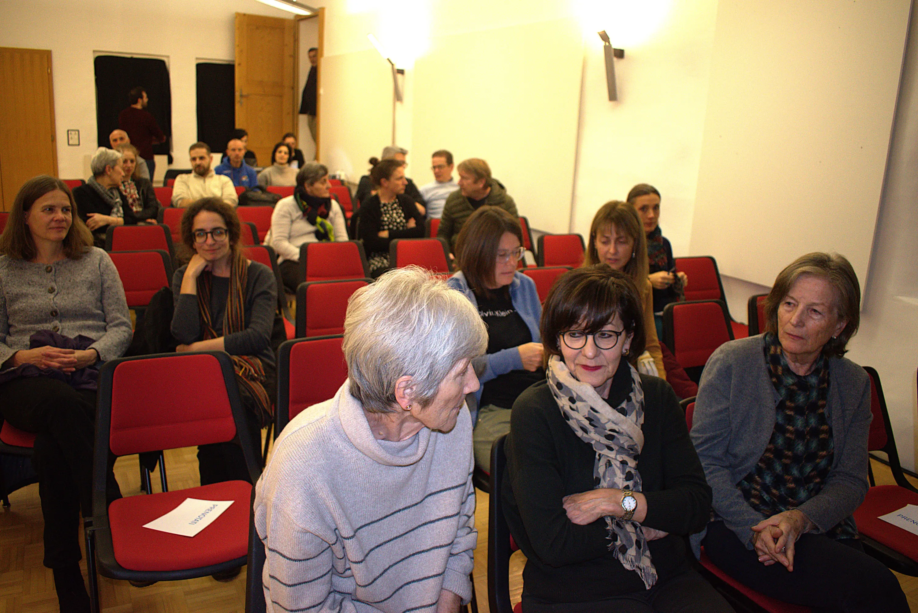 Un gruppo di persone sedute in una sala con sedie rosse, alcune chiacchierano tra loro. Si notano persone di diverse età, alcune sembrano attente e altre sorridenti. Sullo sfondo, altri partecipanti sono visibili.