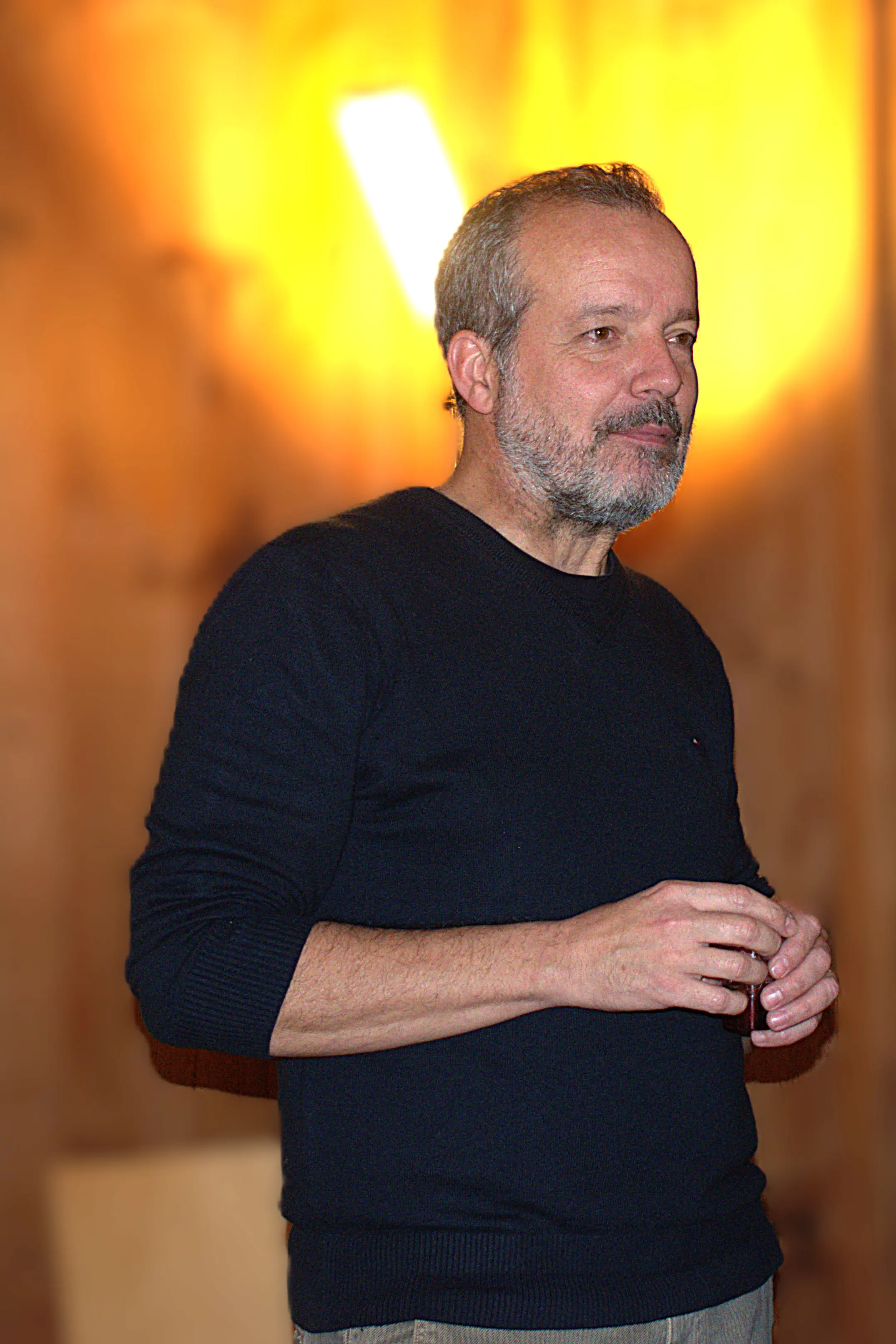 Uomo adulto con barba grigia, indossa un maglione nero e tiene un bicchiere in mano. Sullo sfondo, una luce calda illumina l'ambiente.