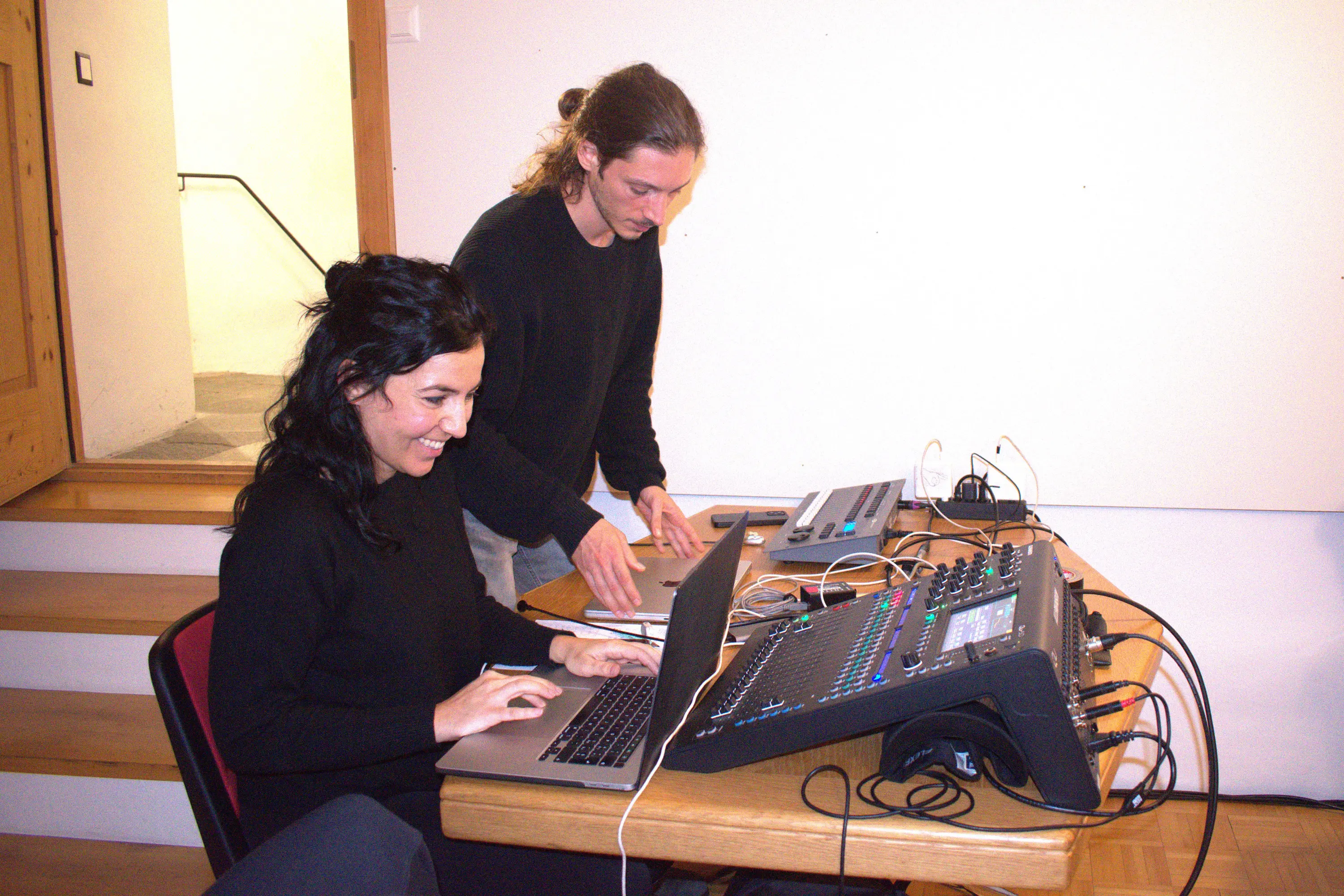 Due persone lavorano a un mixer audio e a un computer portatile. Una donna sta digitando sulla tastiera, mentre un uomo regola i controlli del mixer. Entrambi indossano abbigliamento scuro.