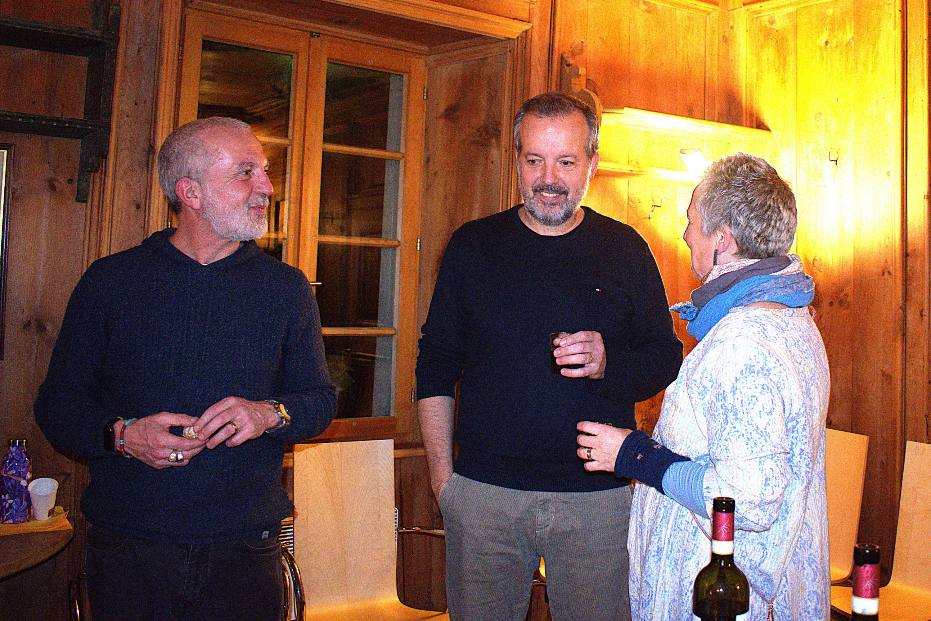 Tre persone si trovano all'interno di una stanza in legno. Due uomini e una donna conversano, sorridendo, con bicchieri in mano. Sul tavolo ci sono alcune bottiglie di vino. La luce calda crea un'atmosfera accogliente.