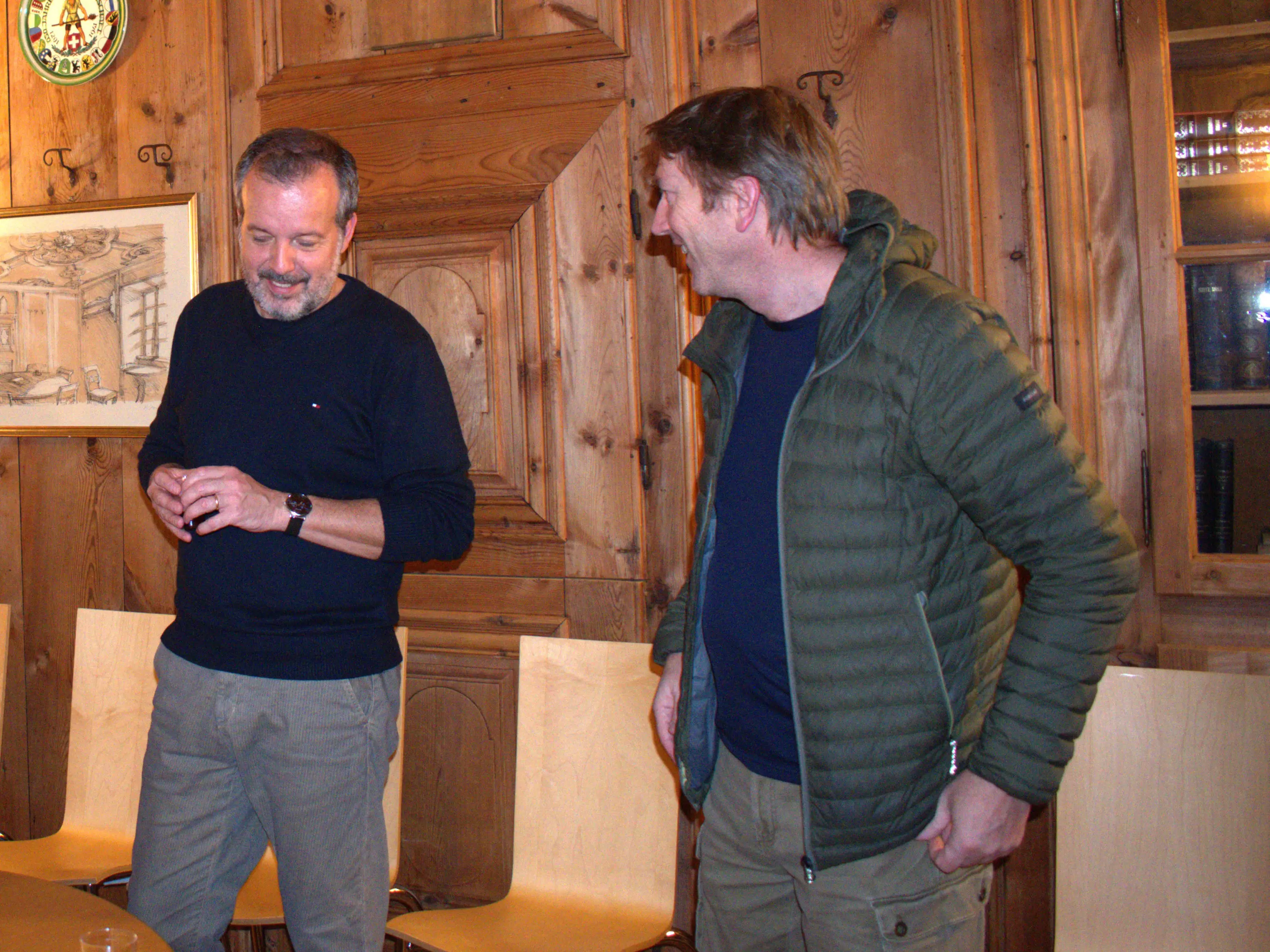 Due uomini sorridenti in una stanza con pareti di legno, uno indossa un maglione scuro e l'altro una giacca leggera. Si scambiano parole in un'atmosfera cordiale.