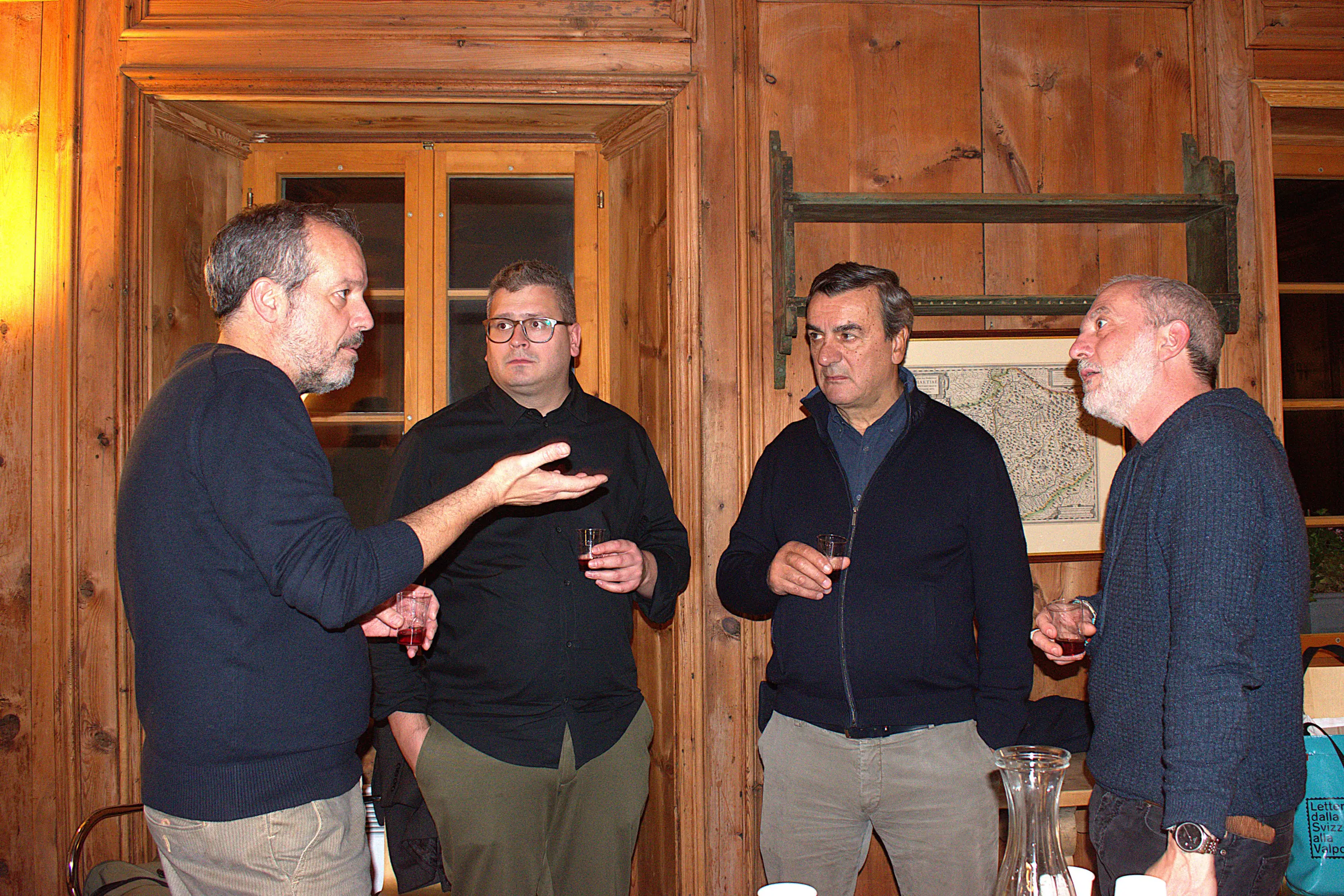 Quattro uomini in conversazione in un ambiente accogliente, con pareti in legno. Due di loro tengono dei bicchieri, mentre uno parla animatamente. L'atmosfera è informale e conviviale.