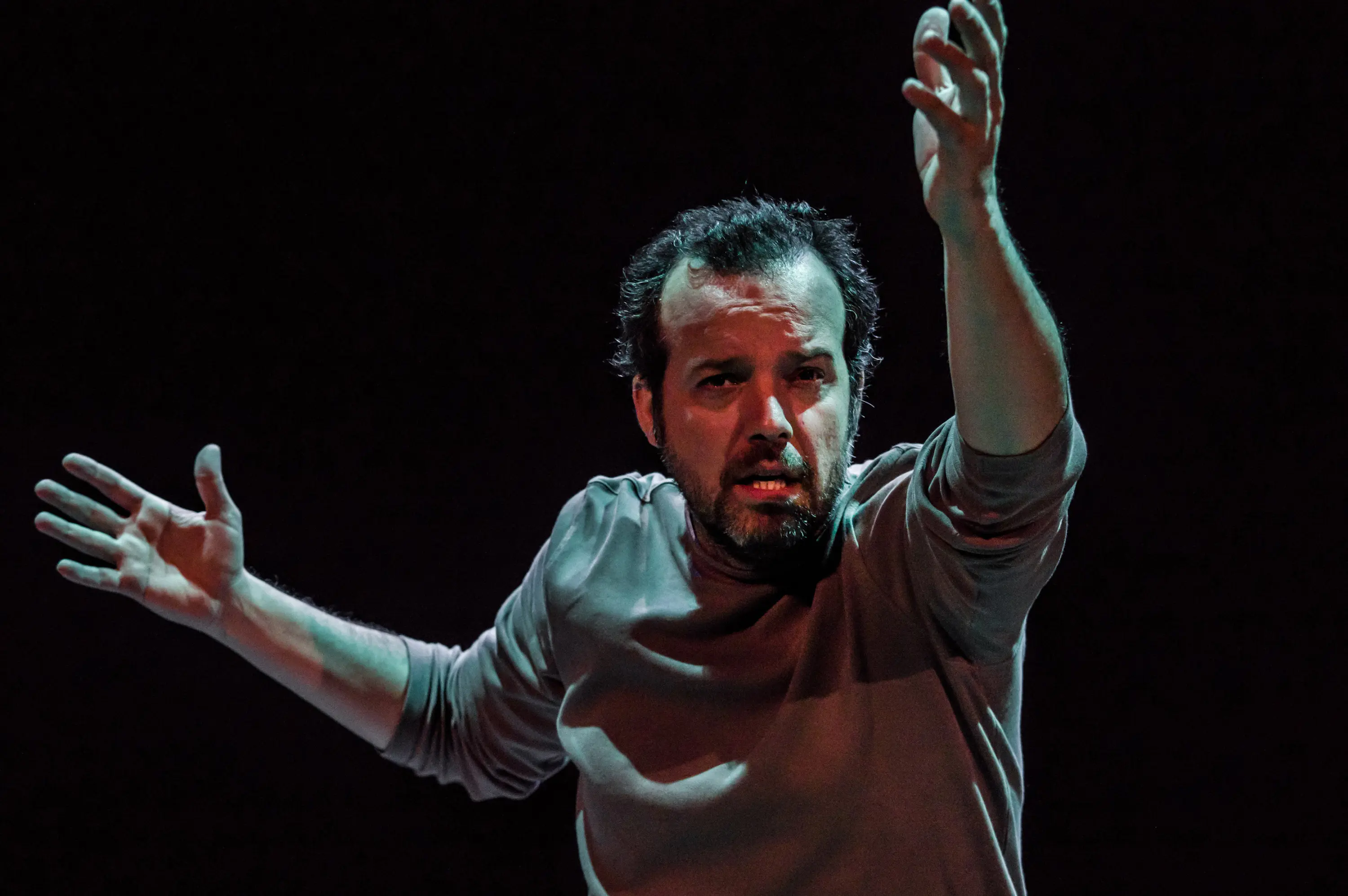 Un uomo si esibisce su un palco buio, con le braccia aperte e un'espressione intensa sul volto, sembra coinvolto in una performance emozionante.