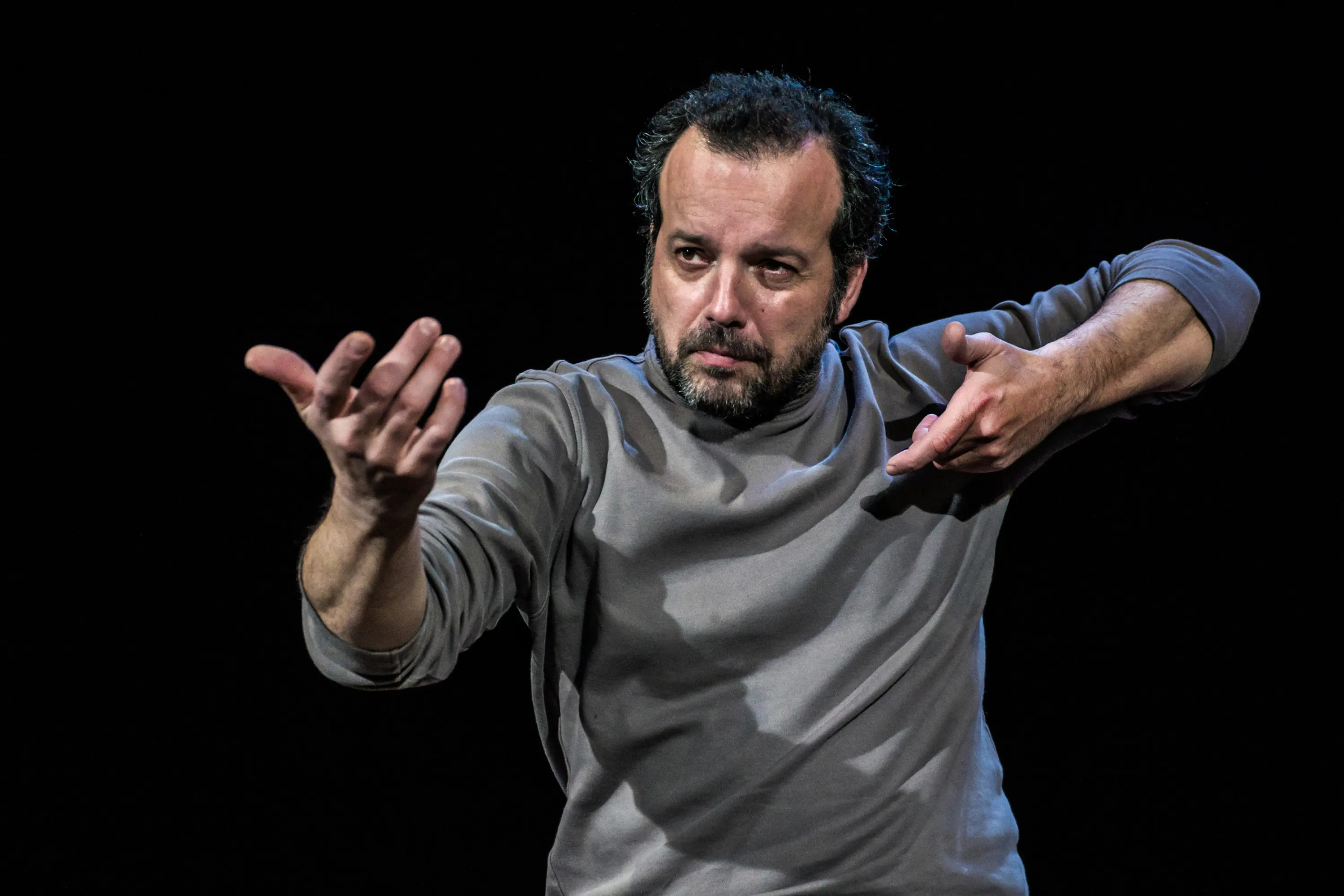 Un uomo si esibisce in un monologo teatrale, vestito con una maglietta grigia scura e con un'espressione intensa. Le sue mani sono in movimento, mentre comunica con passione. Sullo sfondo è presente un'illuminazione scura che mette
