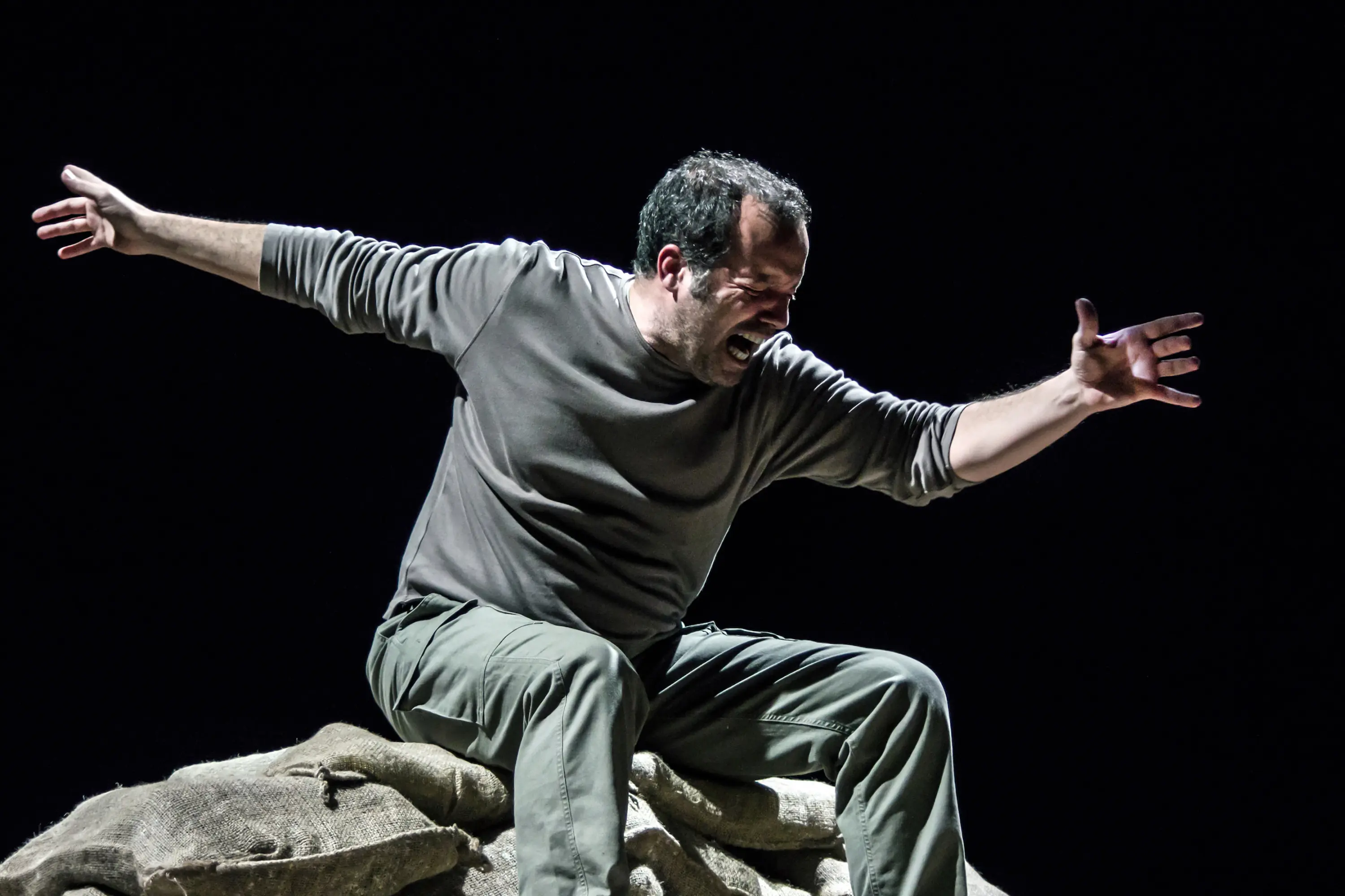 Un uomo si trova sopra un sacco, con le braccia aperte e un'espressione intensa di emozione, mentre sembra esprimere una forte reazione o un urlo. Indossa una maglietta grigia e pantaloni verdi, in un'illuminazione scura