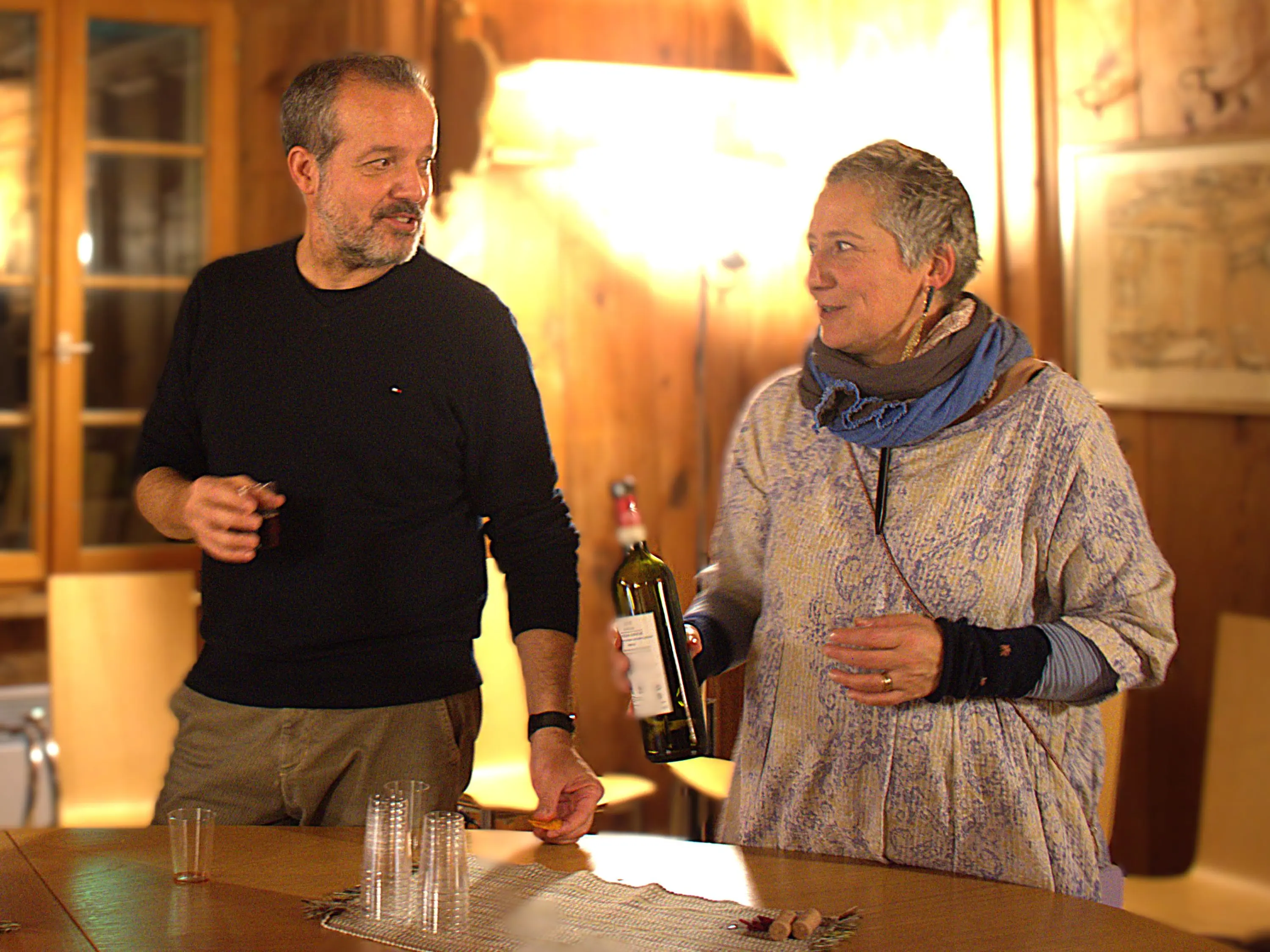 Due persone in una stanza accogliente, sorridendo l'una all'altra. Un uomo con maglione nero tiene un bicchiere, mentre una donna con una sciarpa blu tiene una bottiglia di vino. Luci calde illuminano l'ambiente di legno.