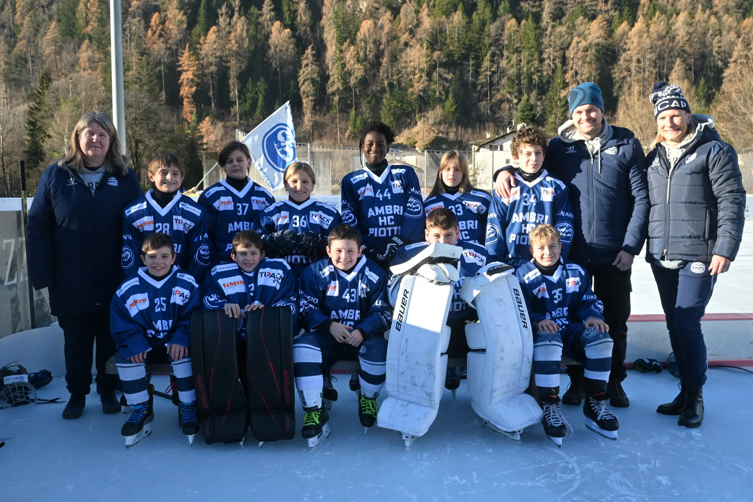 Giovani giocatori di hockey su ghiaccio in divisa blu, sorridenti, in posa con gli allenatori. Sullo sfondo, alberi e montagne. Due portieri in attrezz