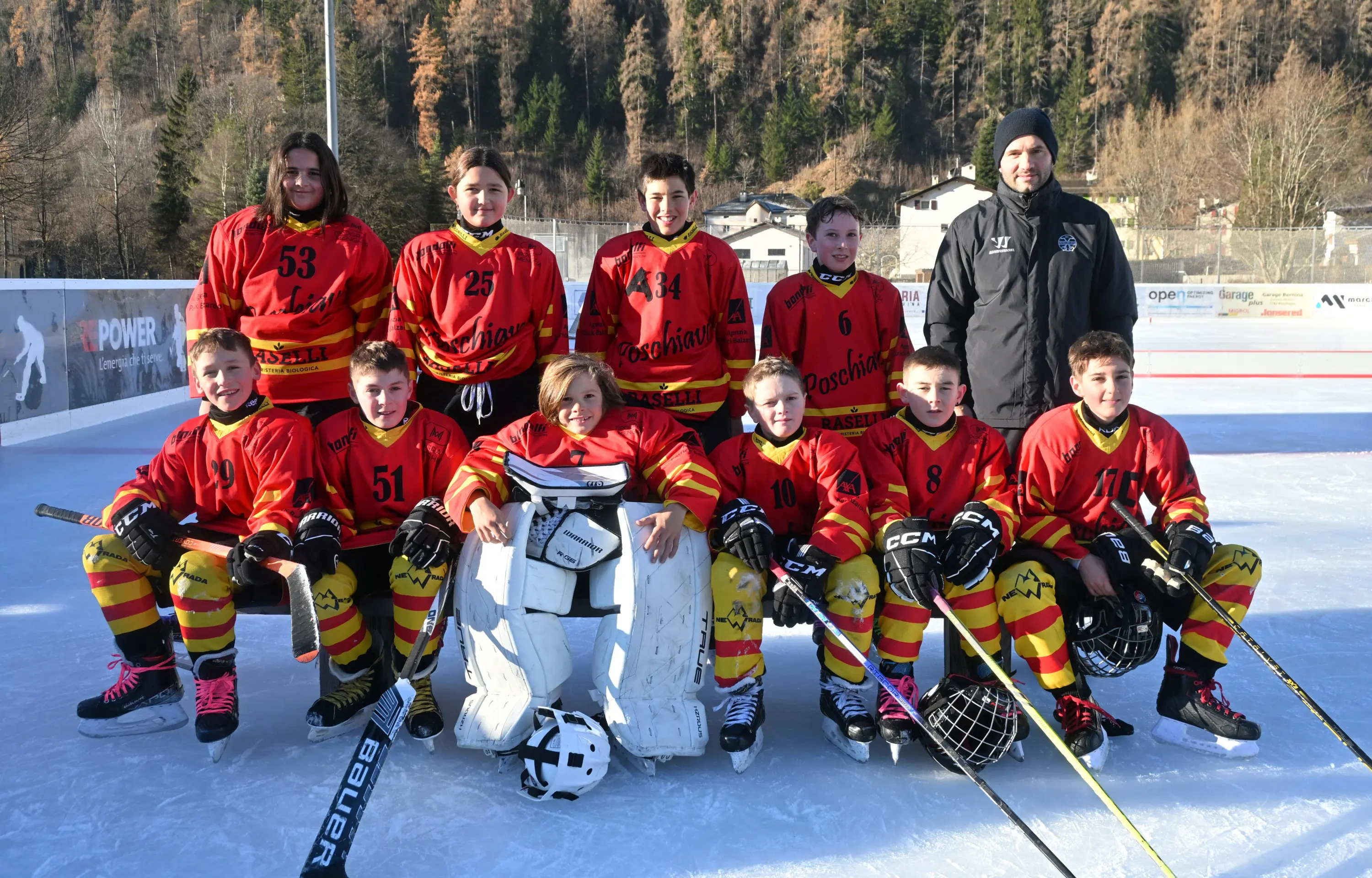 Giocatori di hockey su ghiaccio in uniforme rossa e gialla. Sono in posa sul ghiaccio, con un portiere al centro e un allenatore alla destra. Sullo sf