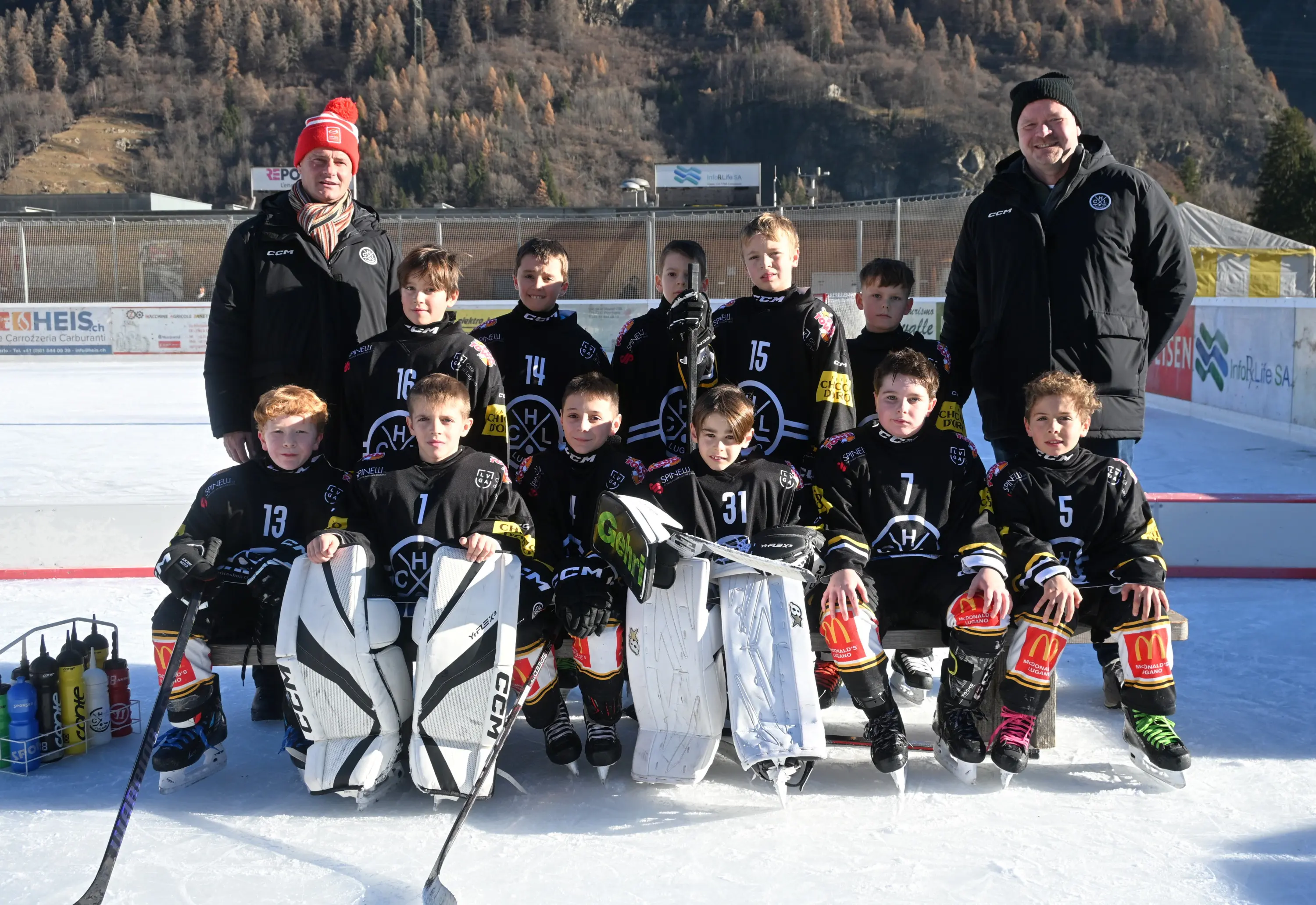 Un gruppo di giovani giocatori di hockey su ghiaccio, indossando divise nere e gialle, posano sul ghiaccio. Due allenatori adults sono in piedi ai lati del gruppo