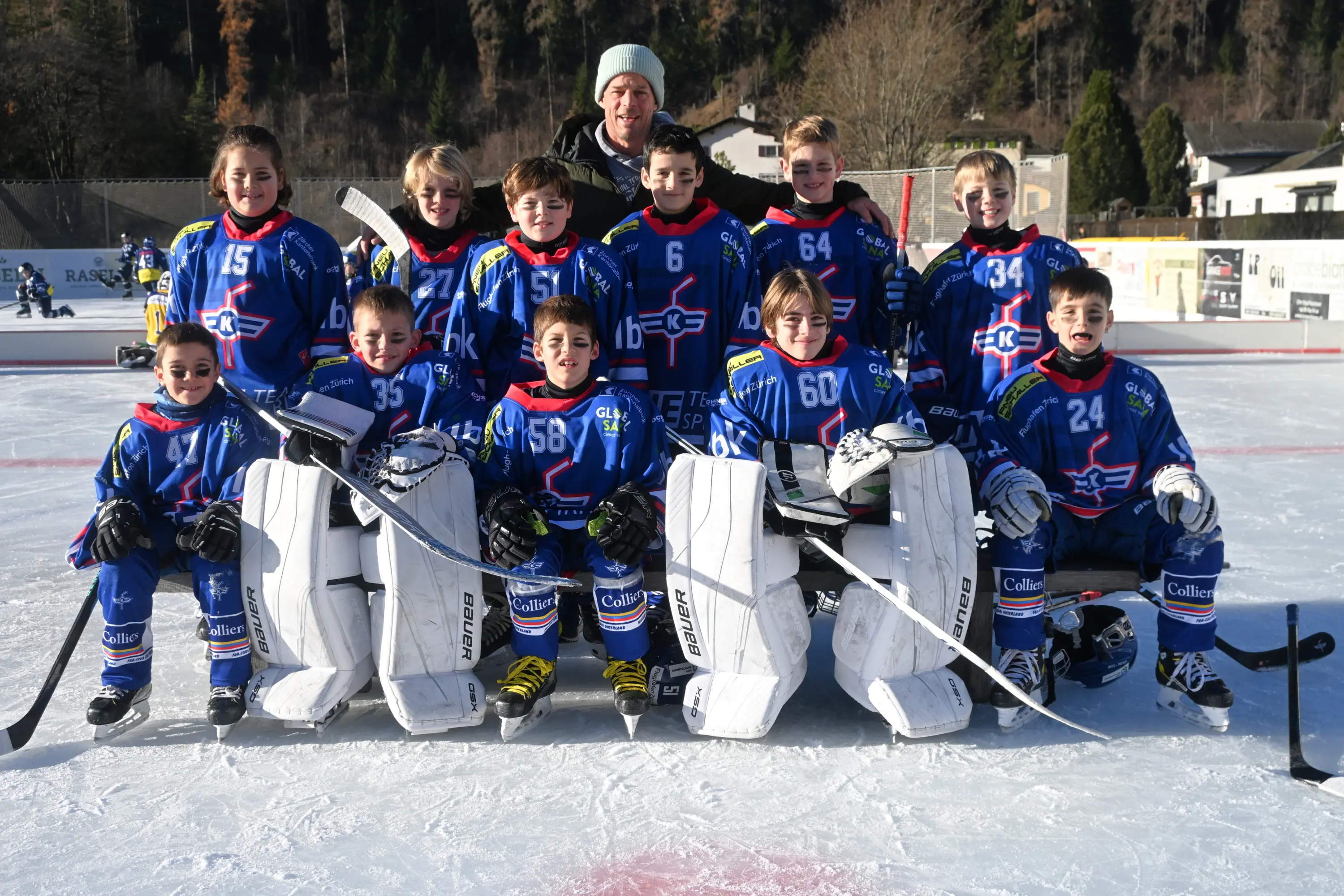 Un gruppo di giovani giocatori di hockey su ghiaccio, in divise blu con il logo della squadra, posano insieme su un campo di ghiaccio. Alcuni indossano maschere