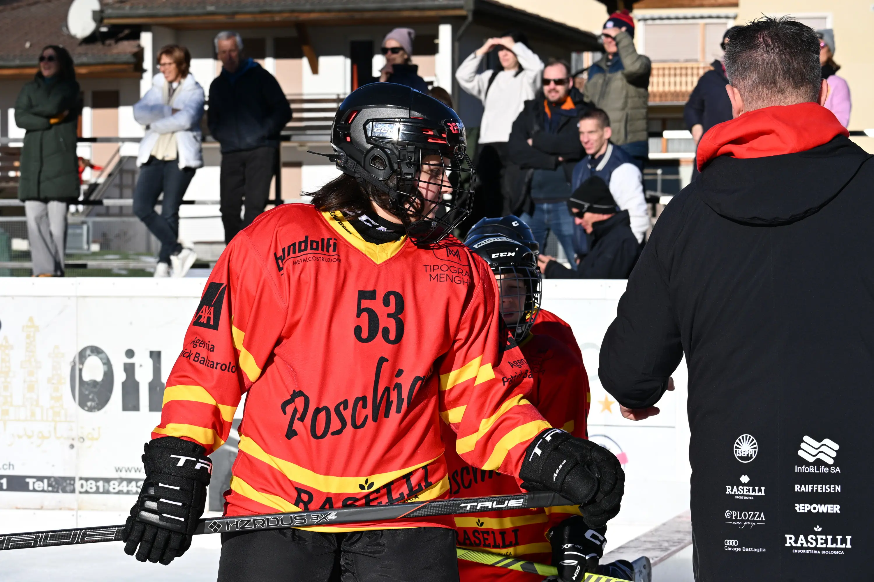 Giocatrice di hockey su ghiaccio in uniforme rossa e gialla, numero 53, interagisce con un allenatore sul campo. Sullo sfondo, alcune persone assistono alla