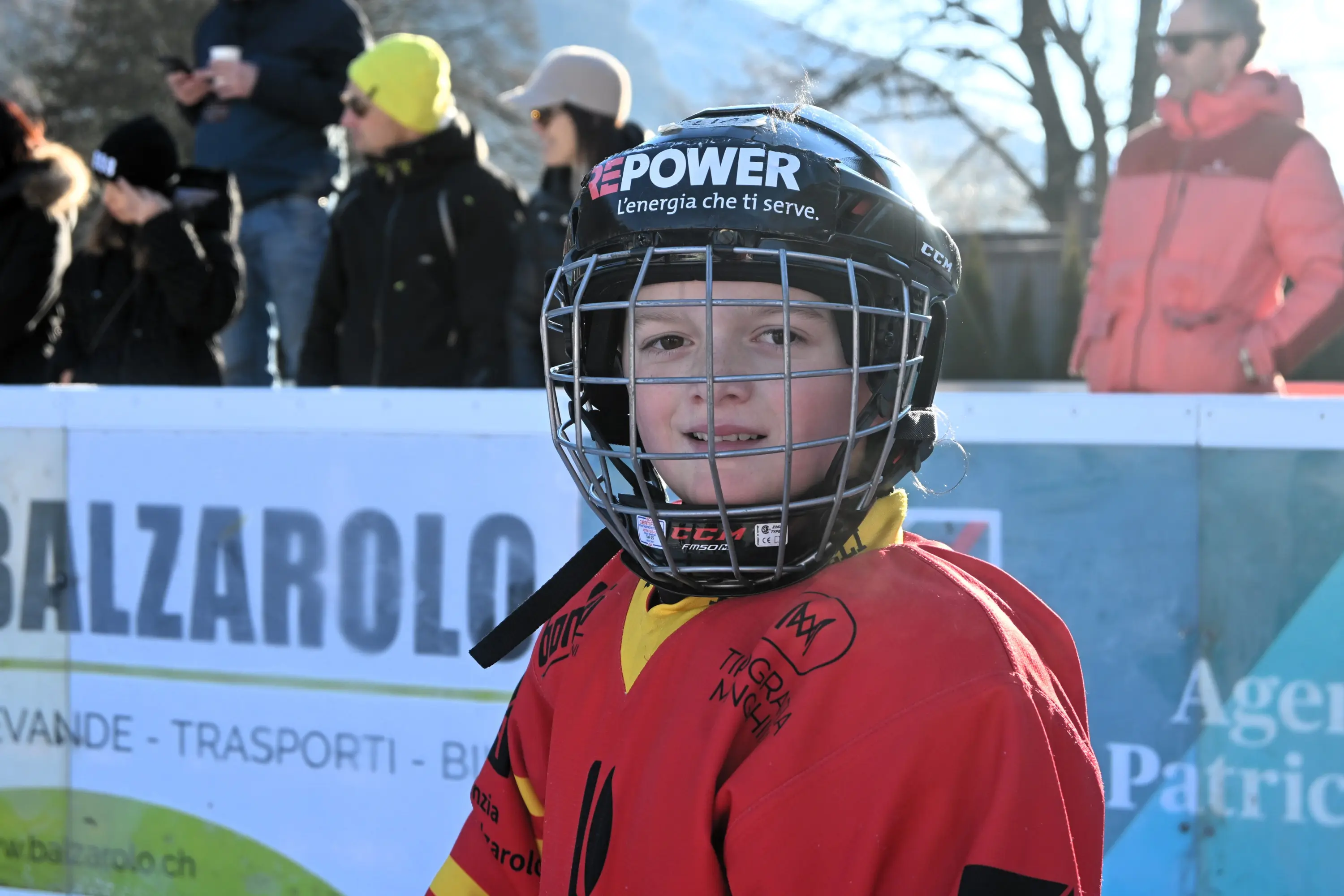 Giovane giocatore di hockey su ghiaccio con caschetto, sorridente e vestito con una maglia rossa e gialla, in un ambiente all'aperto con pubblico sullo sf