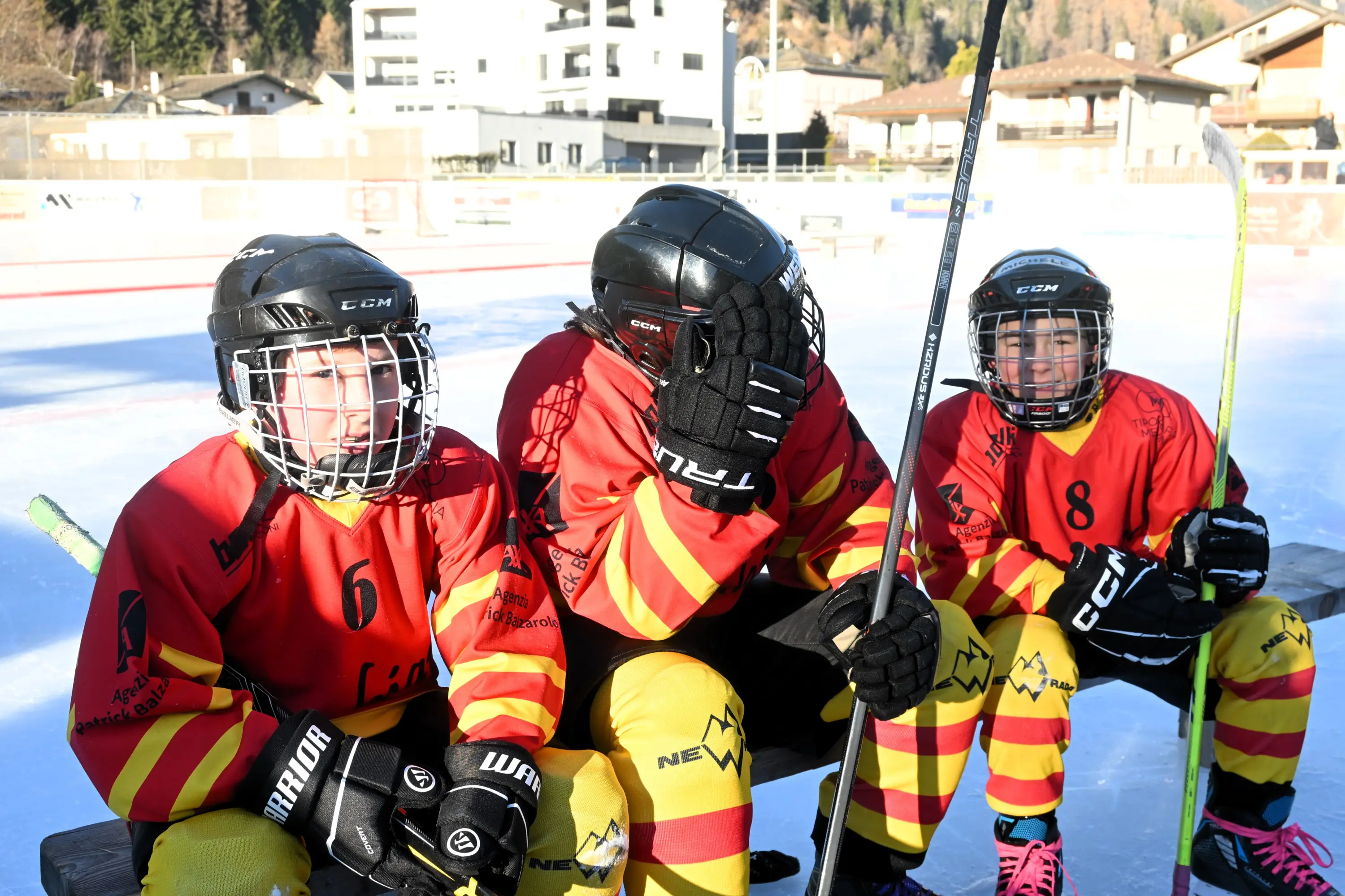 Tre giovani giocatori di hockey su ghiaccio seduti su una panchina. Indossano maglie rosse e gialle con numeri, caschi neri e guanti.