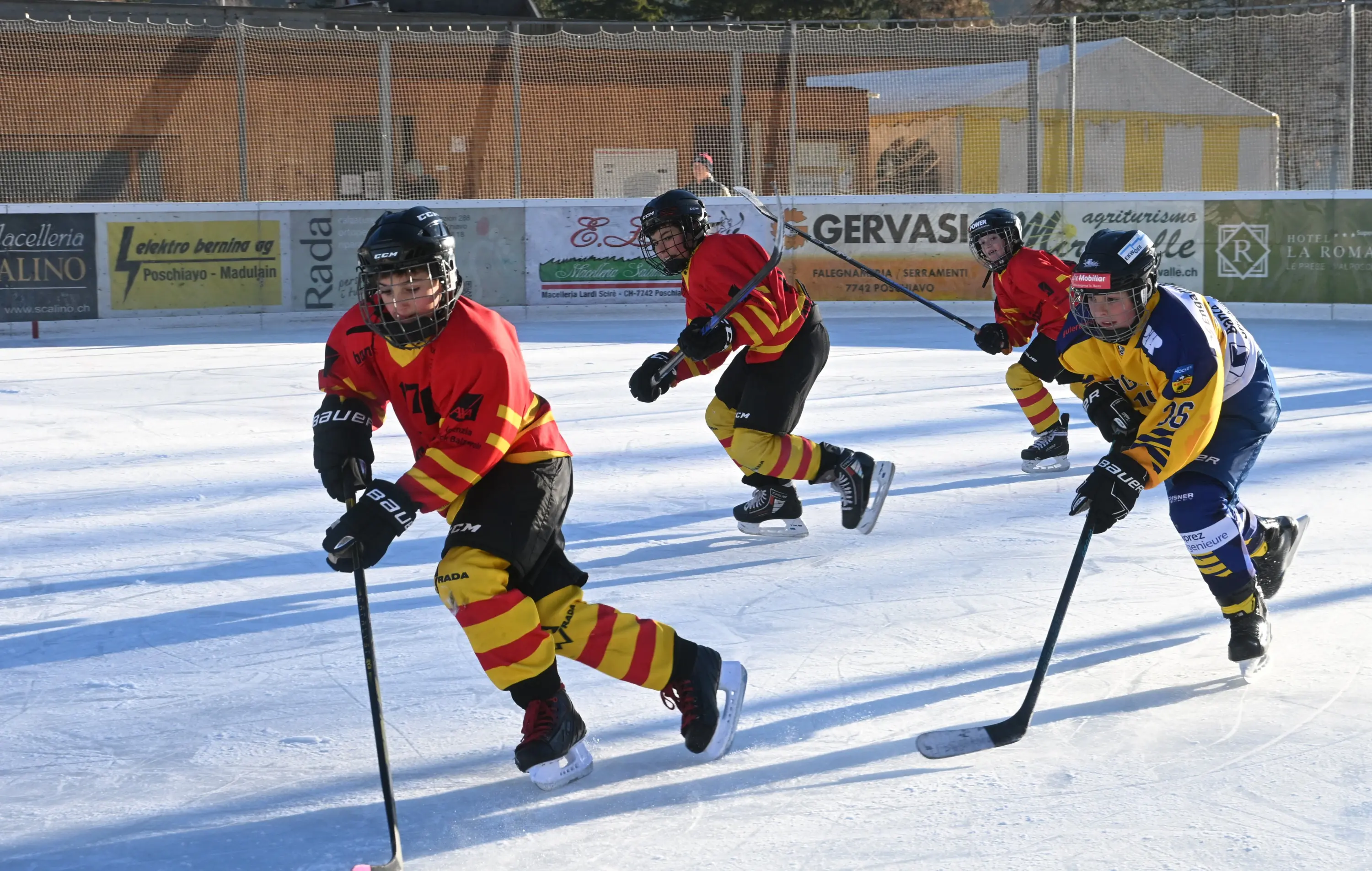 Giocatori di hockey su ghiaccio in azione, indossando maglie rosse e gialle, mentre patinano su un campo di ghiaccio. Due giocatori si trov
