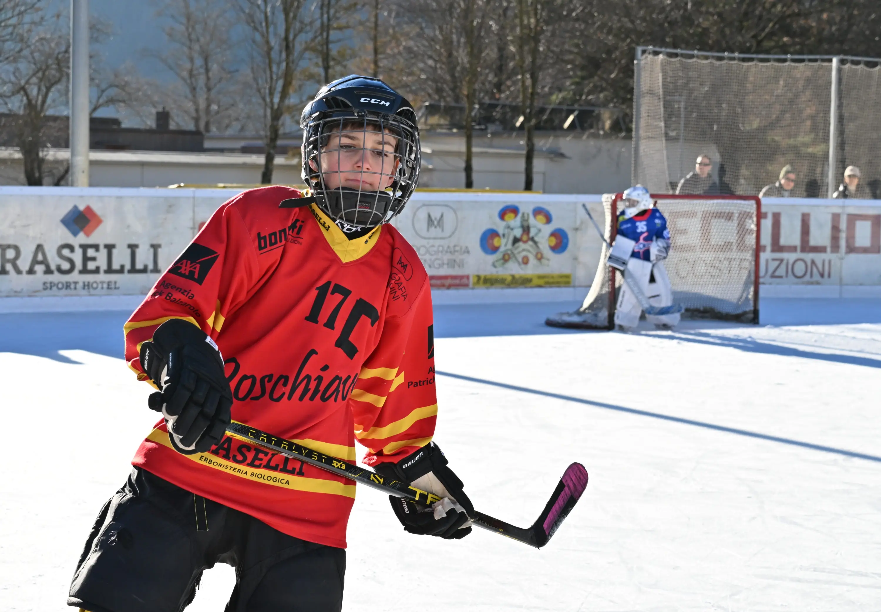 Giocatore di hockey su ghiaccio in azione, indossa una maglia rossa e gialla con un casco protettivo. Sullo sfondo, un portiere in uniforme blu
