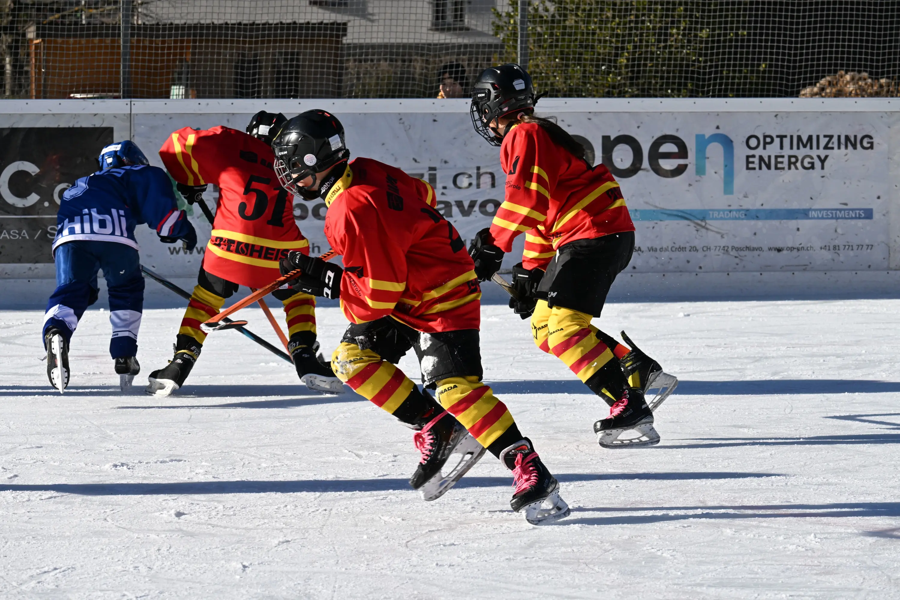 Giocatori di hockey su ghiaccio in azione, indossando maglie rosse e gialle. Si vedono due giocatori in movimento, mentre un terzo si prepara a