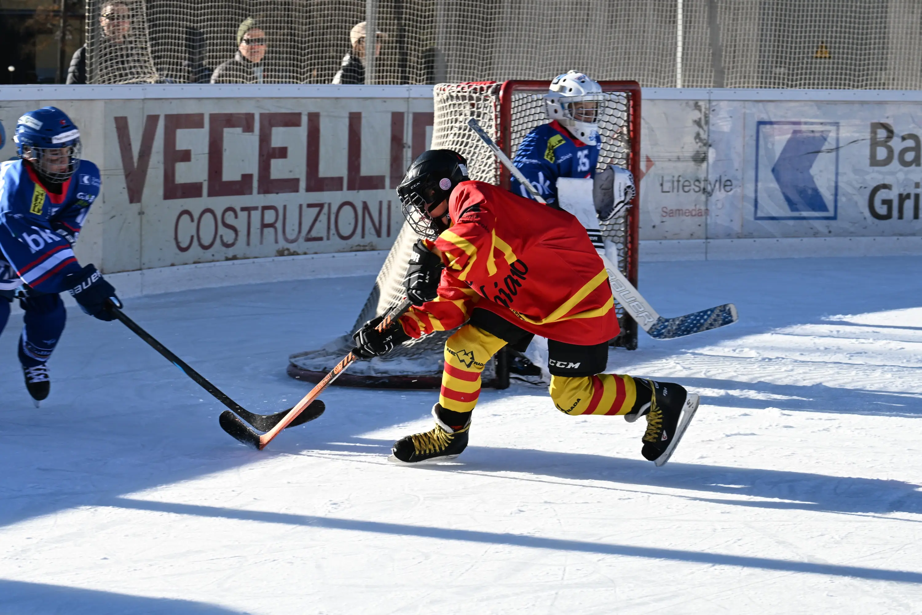 Giocatore di hockey su ghiaccio in maglia rossa e gialla mentre colpisce il puck. Sullo sfondo, un portiere e un altro giocatore avverso.