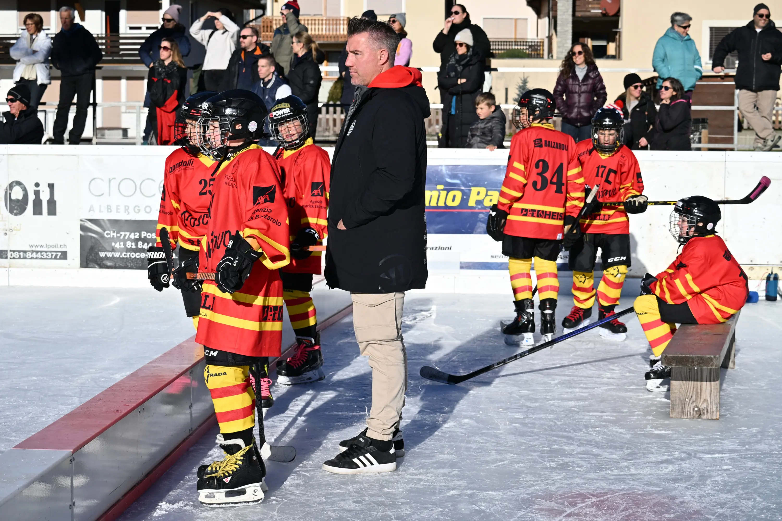Un allenatore osserva i giovani giocatori di hockey su ghiaccio, indossando maglie rosse e gialle, mentre alcuni sono seduti su una panchina e altri si