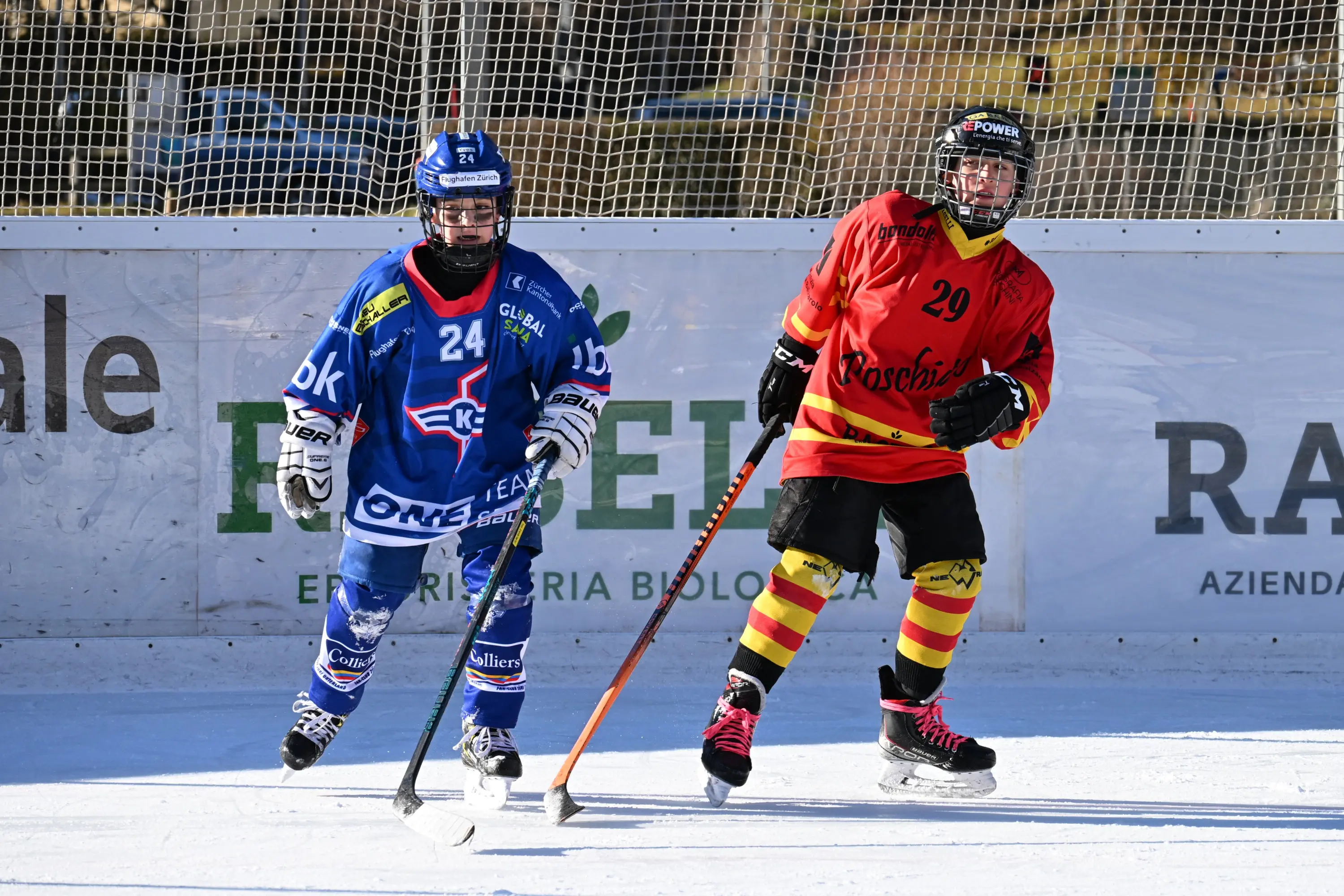 Due giocatori di hockey su ghiaccio in azione. Uno indossa una maglia blu con numero 24, l'altro una maglia rossa e gialla con numero 29.