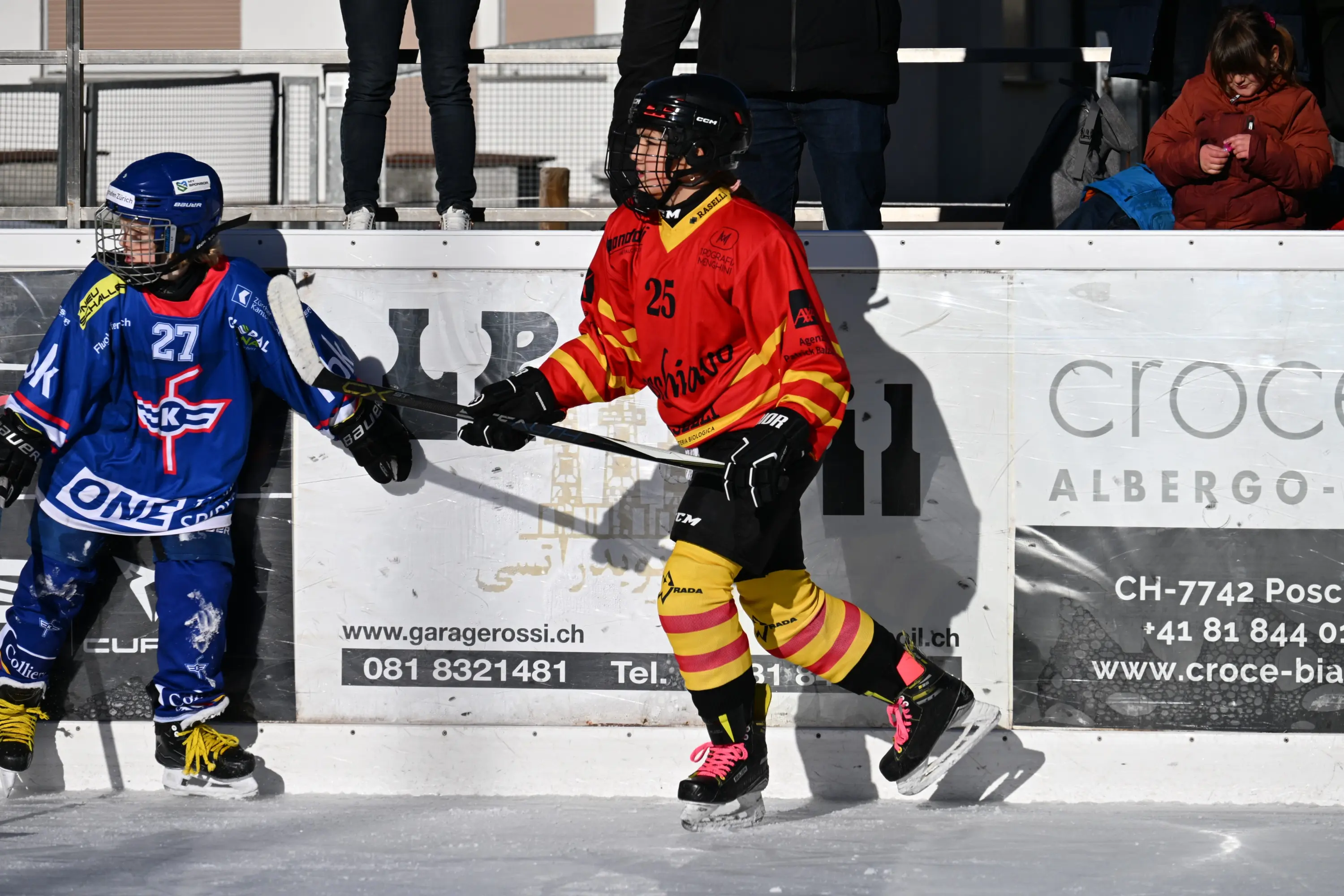 Due giovani giocatori di hockey su ghiaccio in azione sul campo. Uno indossa una divisa blu e l'altro una divisa rossa e gialla, entrambi con caschi
