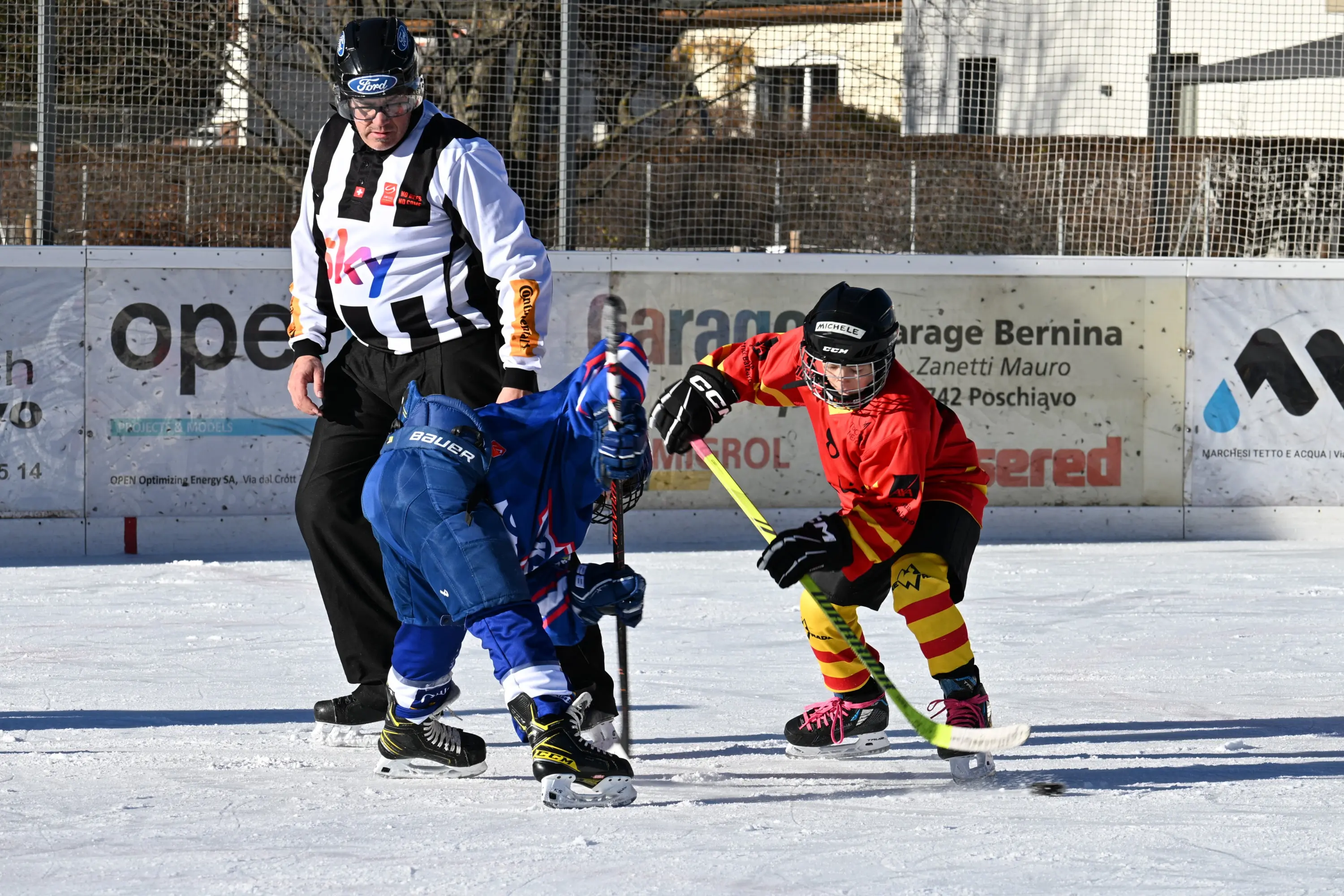 Due giovani giocatori di hockey su ghiaccio stanno combattendo per il disco, mentre un arbitro li osserva. Un giocatore indossa una divisa blu, l'altro una maglia