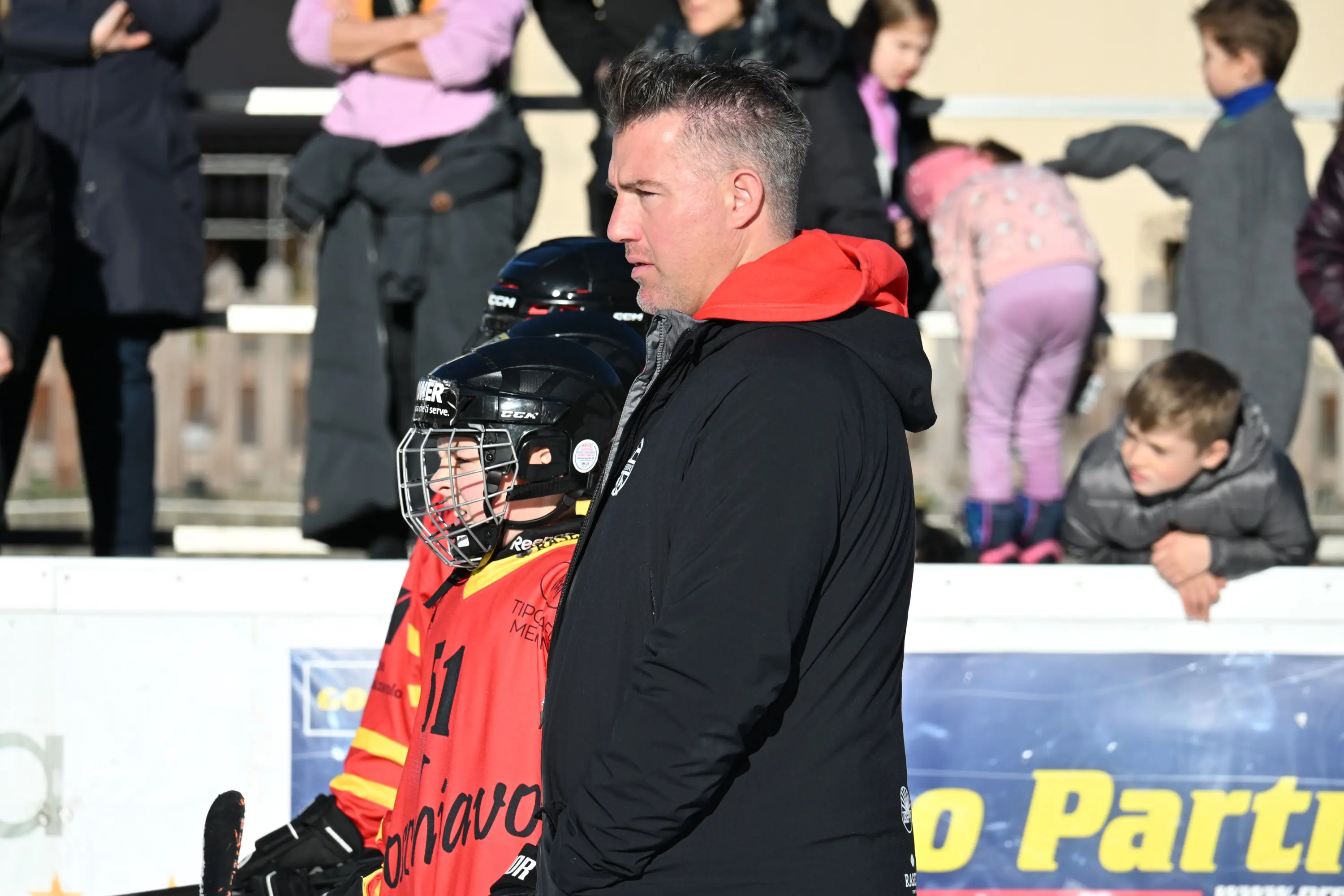 Un allenatore osserva attentamente un giovane giocatore di hockey su ghiaccio, mentre altri bambini si divertono sullo sfondo.