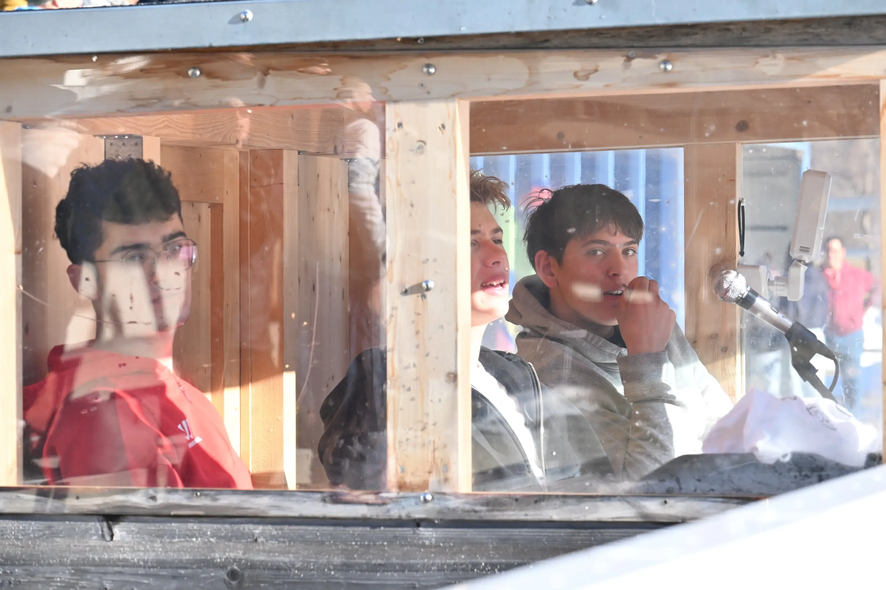 Tre ragazzi seduti all'interno di una struttura di legno, sembra essere un banco di lavoro. Le pareti sono trasparenti e si vedono riflessi esterni. I ragazzi