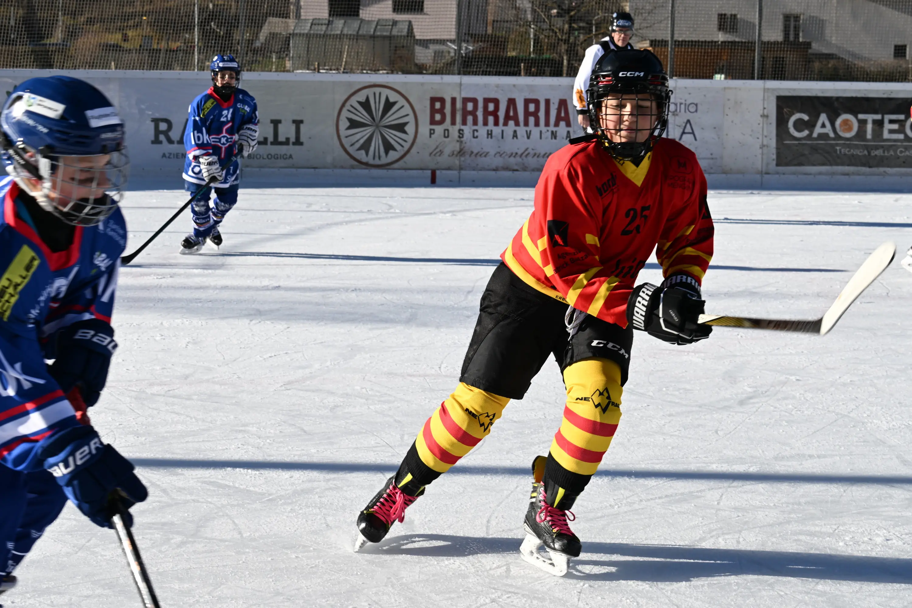 Giocatore di hockey su ghiaccio in azione, indossando una maglia rossa e nera con strisce gialle. Altri giocatori sono visibili sul ghiaccio