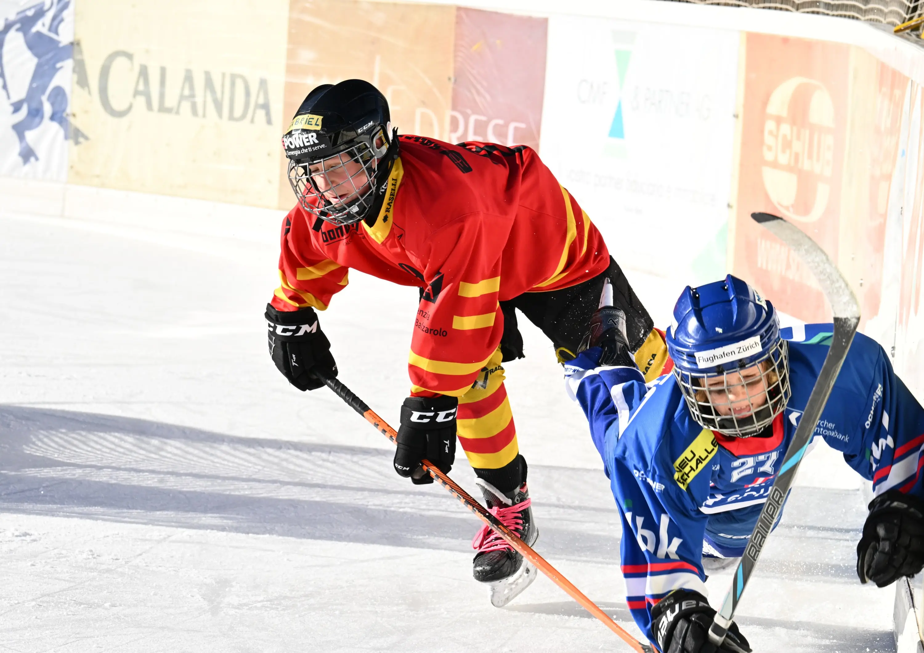 Due giocatori di hockey su ghiaccio si sfidano su un campo ghiacciato. Uno indossa una maglietta rossa con strisce gialle mentre l'altro indossa