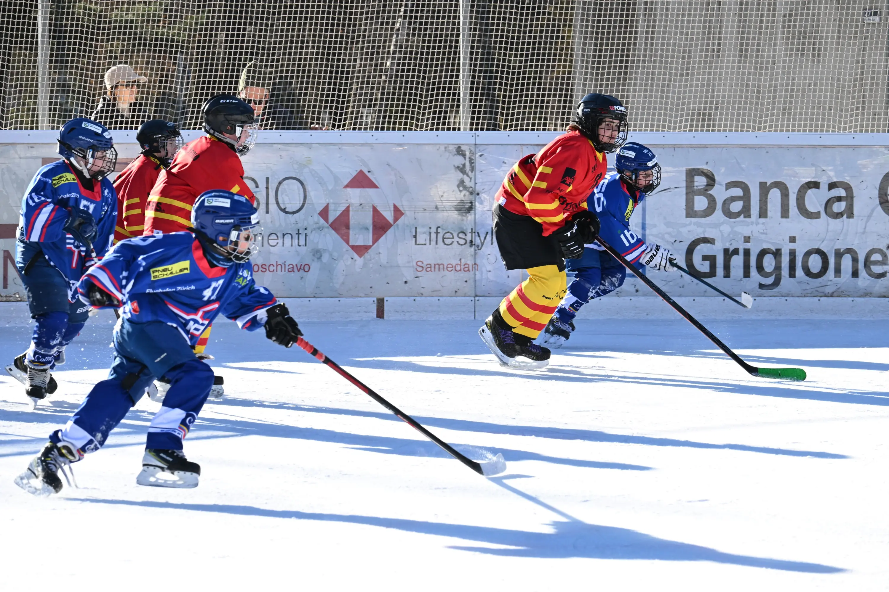 Giocatori di hockey su ghiaccio in azione, alcuni indossano maglie rosse e altri blu. Si trovano su un campo all'aperto, con bande di protezione e