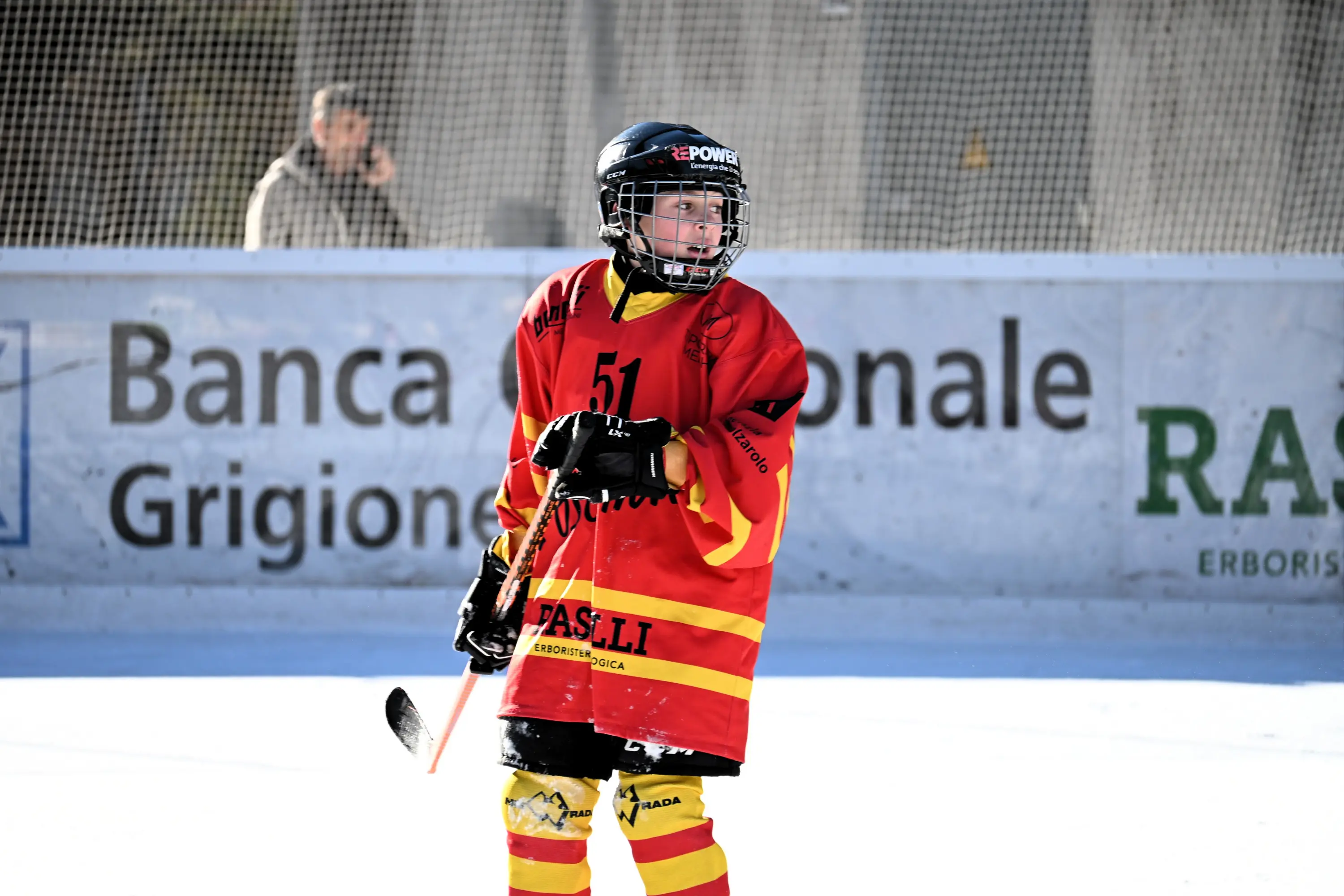 Giovane giocatore di hockey su ghiaccio in una divisa rossa e gialla, con un bastone in mano, in un campo di ghiaccio all'aperto. Sullo sfondo