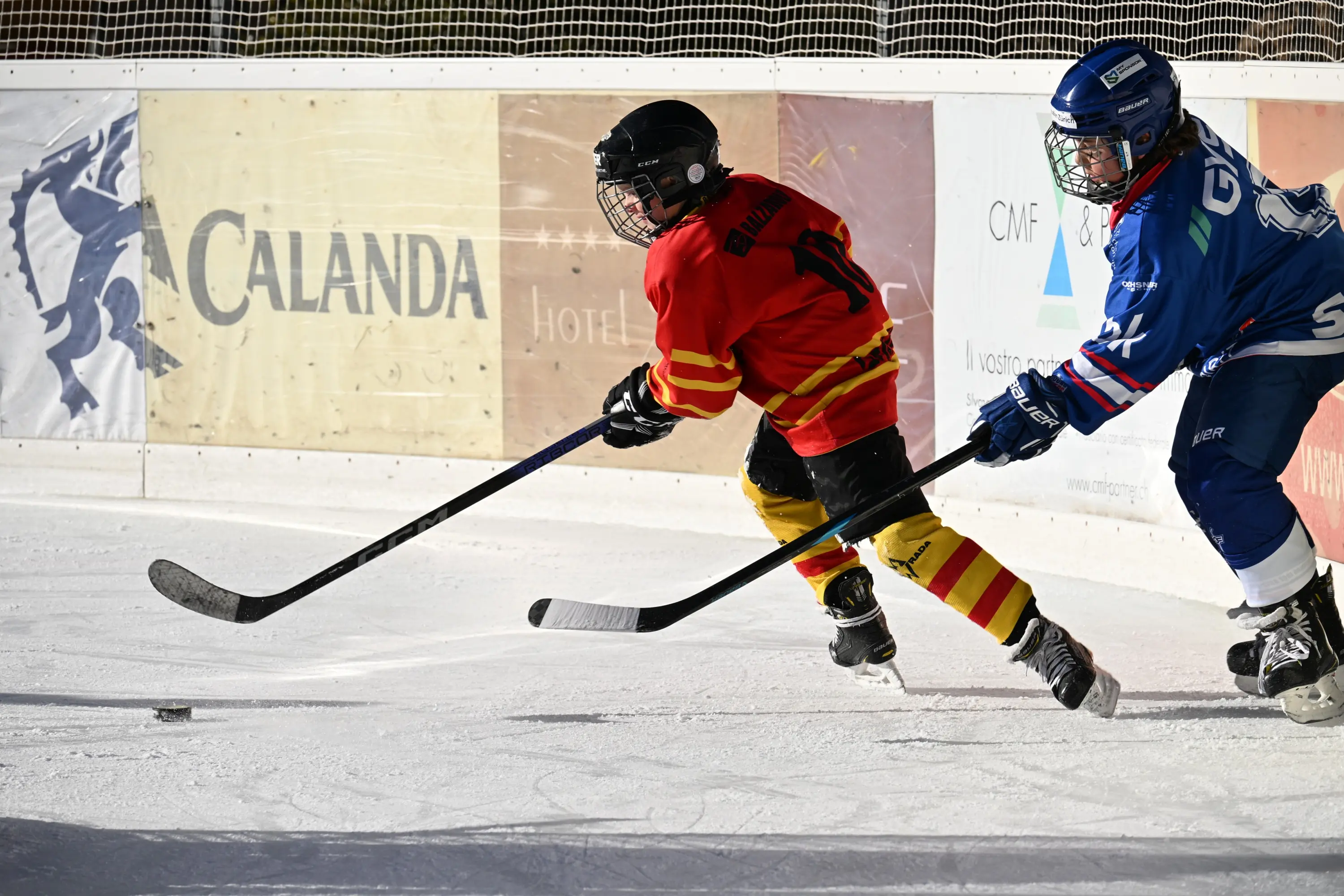 Due giocatori di hockey su ghiaccio in azione. Uno indossa una maglia rossa con strisce gialle e si dirige verso il disco, mentre l’altro, vestito di