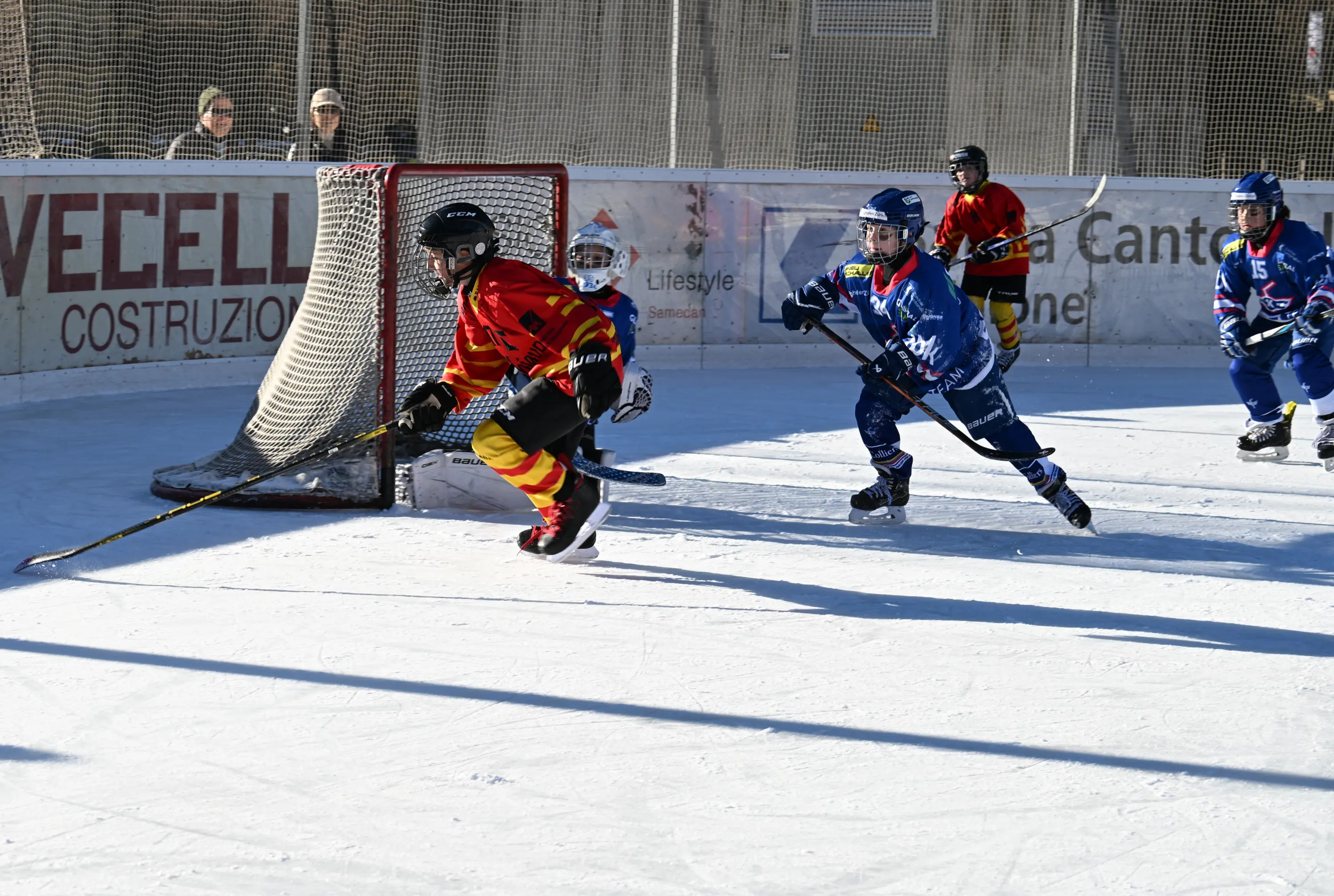 Giocatori di hockey su ghiaccio in azione, uno in maglia rossa e gialla che controlla il disco, mentre un altro in maglia blu lo insegue. Sullo