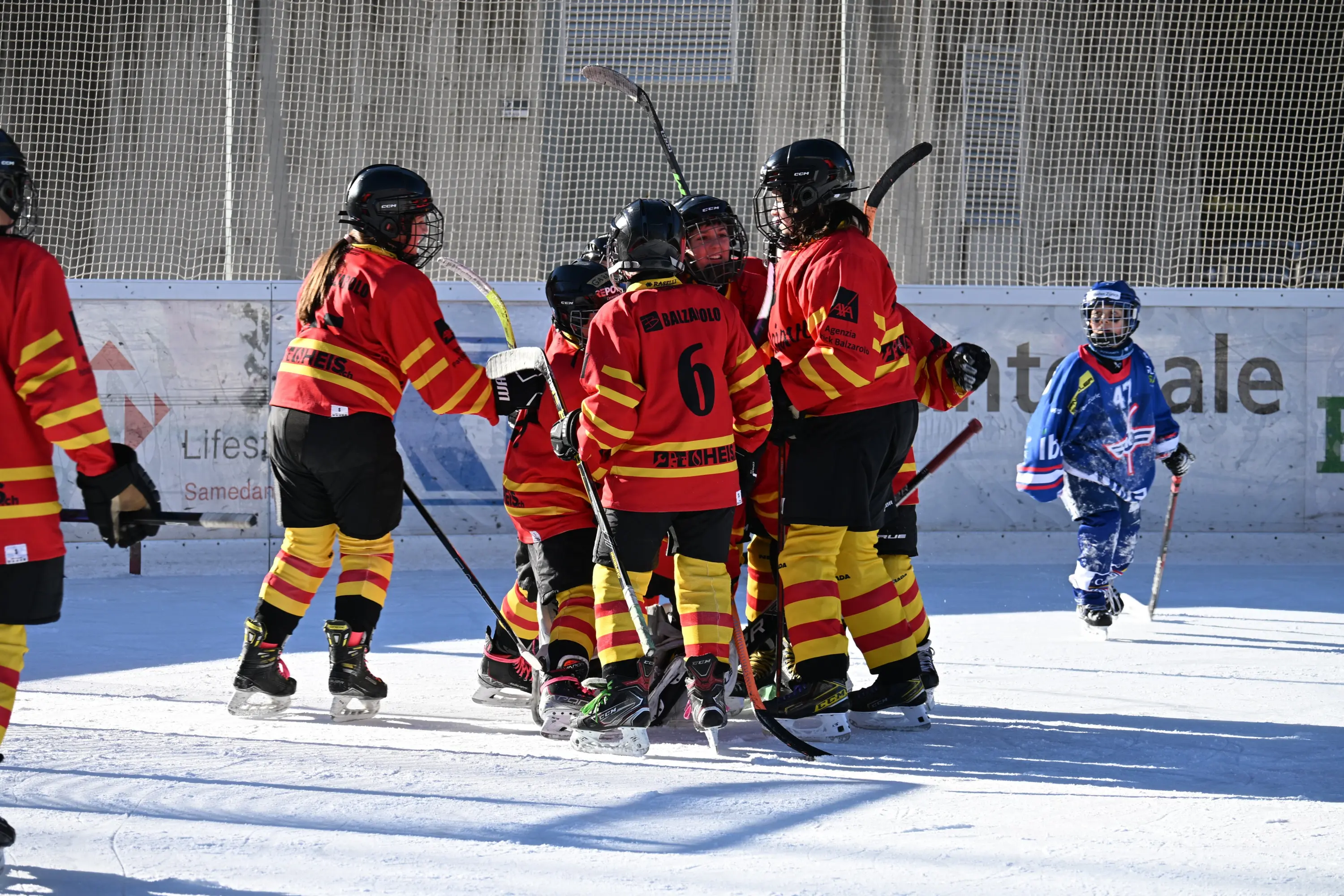 Giocatori di hockey su ghiaccio festeggiano un gol, indossando divise rosse e gialle. Un altro giocatore, in uniforme blu, osserva.