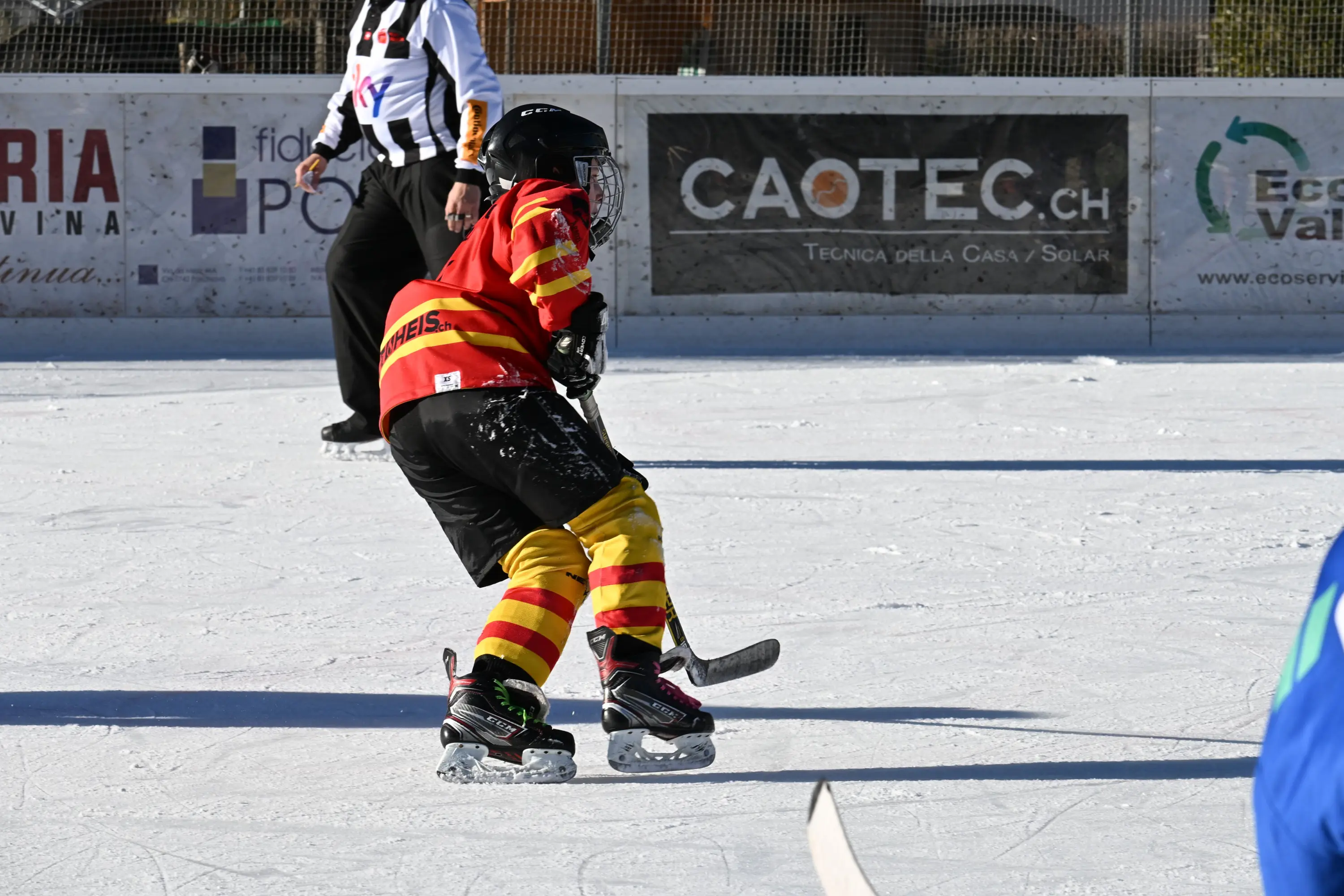 Giocatore di hockey su ghiaccio con maglia rossa e strisce gialle, in movimento sul ghiaccio. Sullo sfondo, un arbitro e pannelli pubblicitari.
