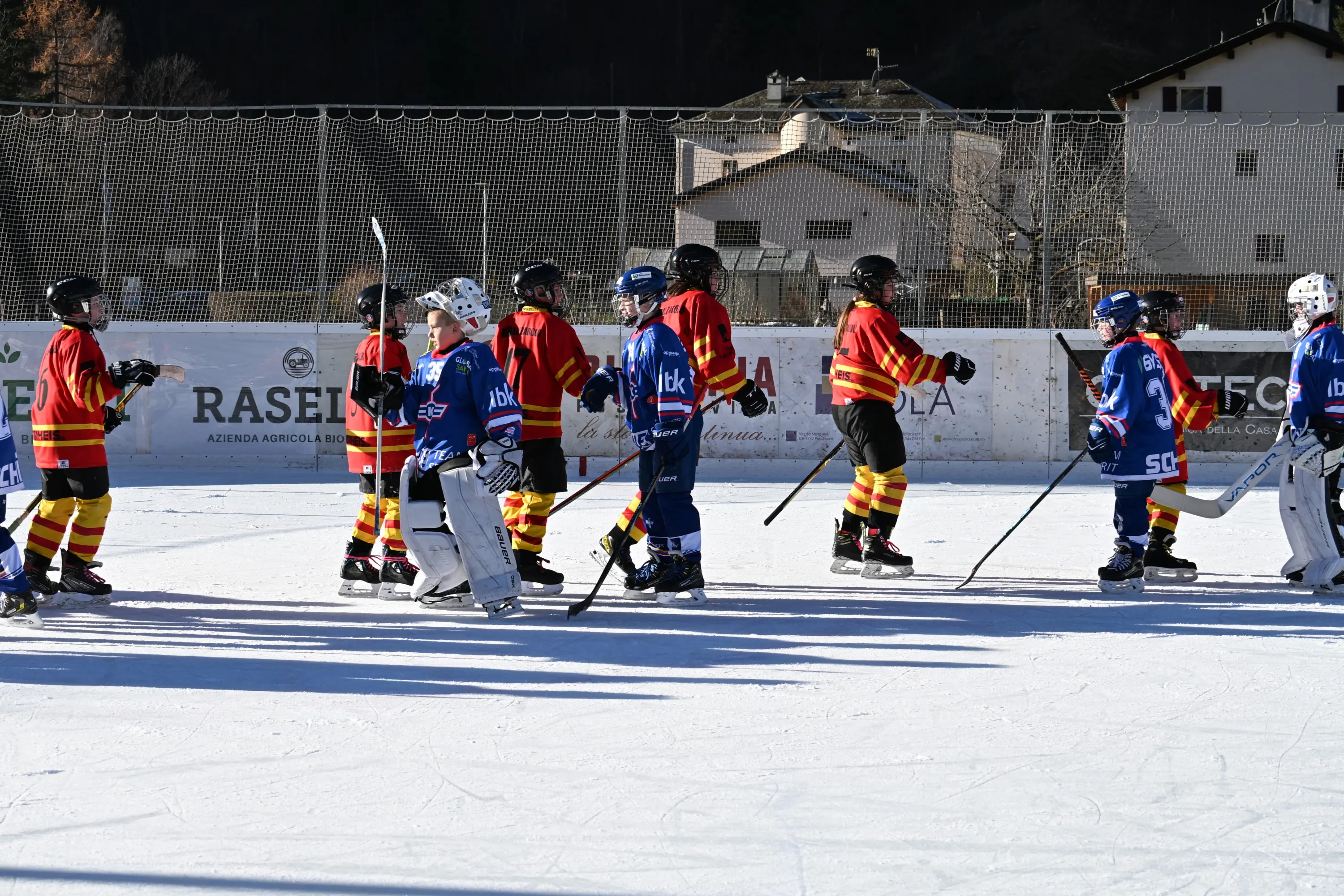 Giovani giocatori di hockey su ghiaccio in un campo all'aperto. Indossano divise rosse e blu mentre si salutano dopo una partita. Sullo sfondo, edifici