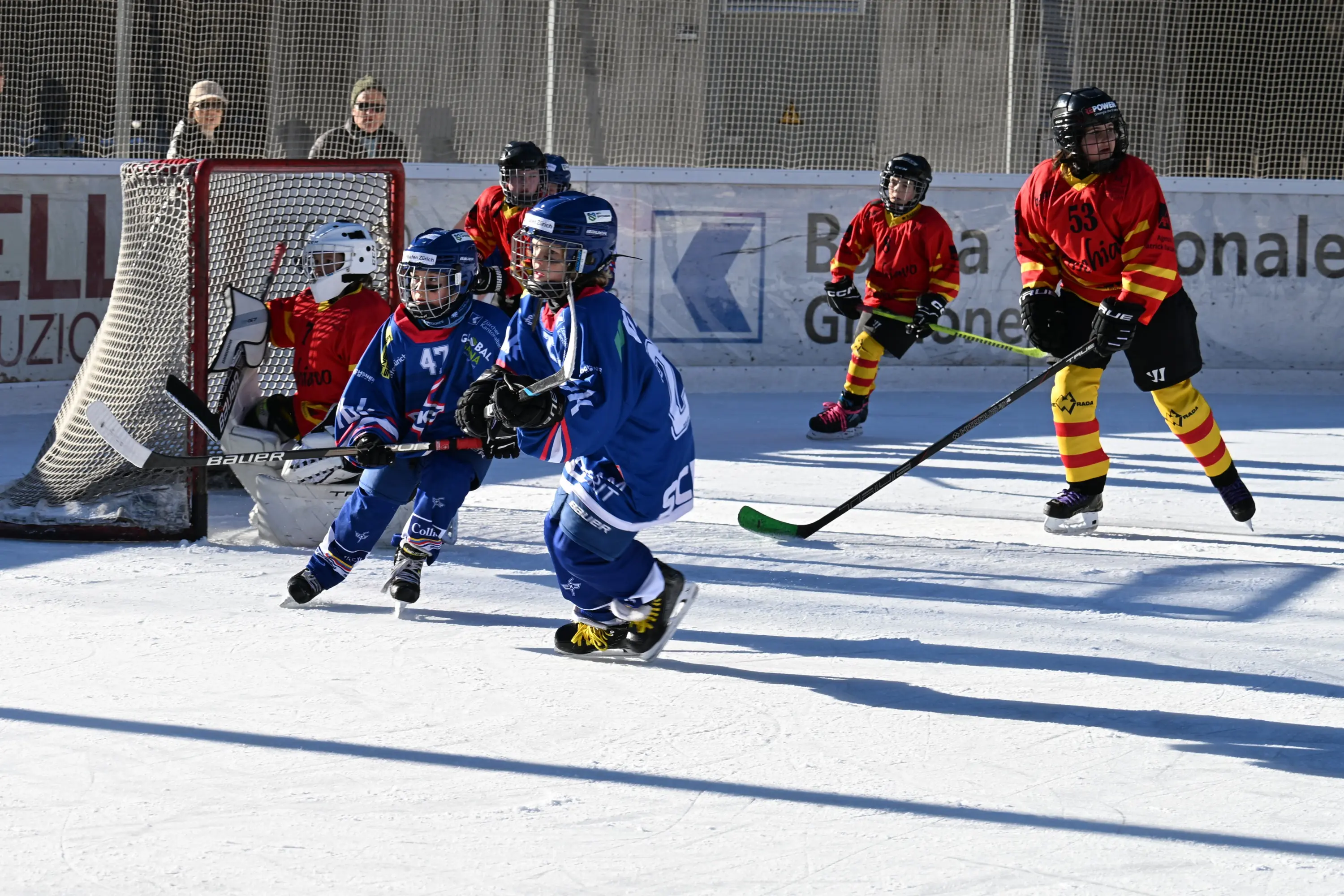 Due squadre di hockey su ghiaccio in azione. Un giocatore in maglia blu si muove verso la porta, mentre due avversari in maglia rossa e gialla si