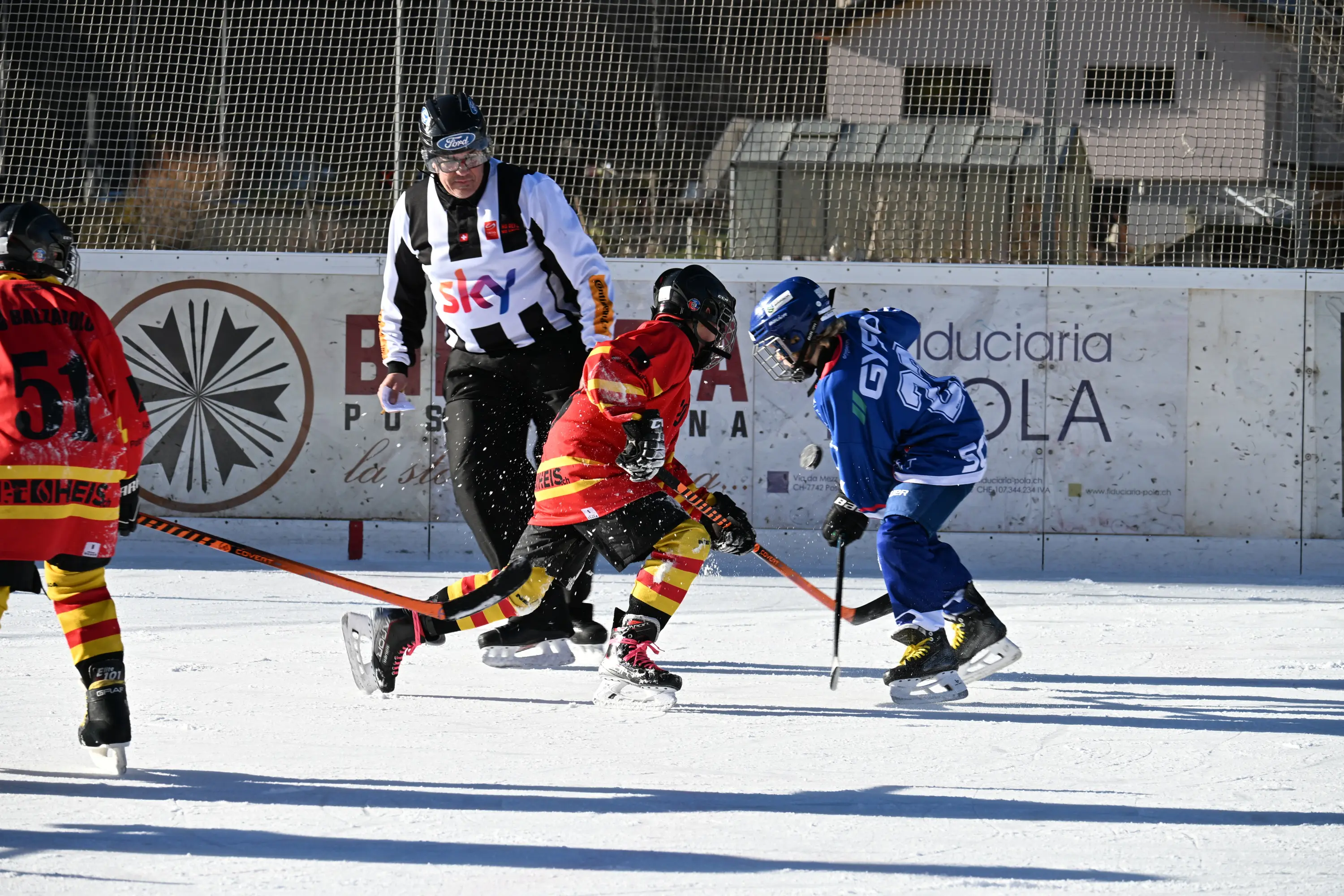 Giocatori di hockey su ghiaccio in azione, con uno in uniforme rossa e gialla che cerca di superare un avversario in blu. Un arbitro osserva la scena