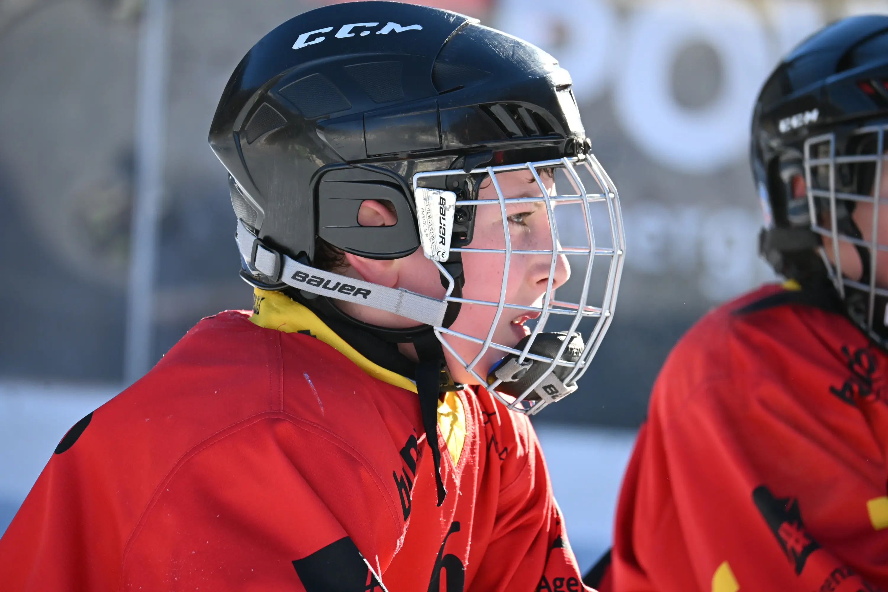 Giovane giocatore di hockey su ghiaccio in attesa di entrare in campo, indossa un casco nero, una visiera e una maglietta rossa. Il suo sguardo