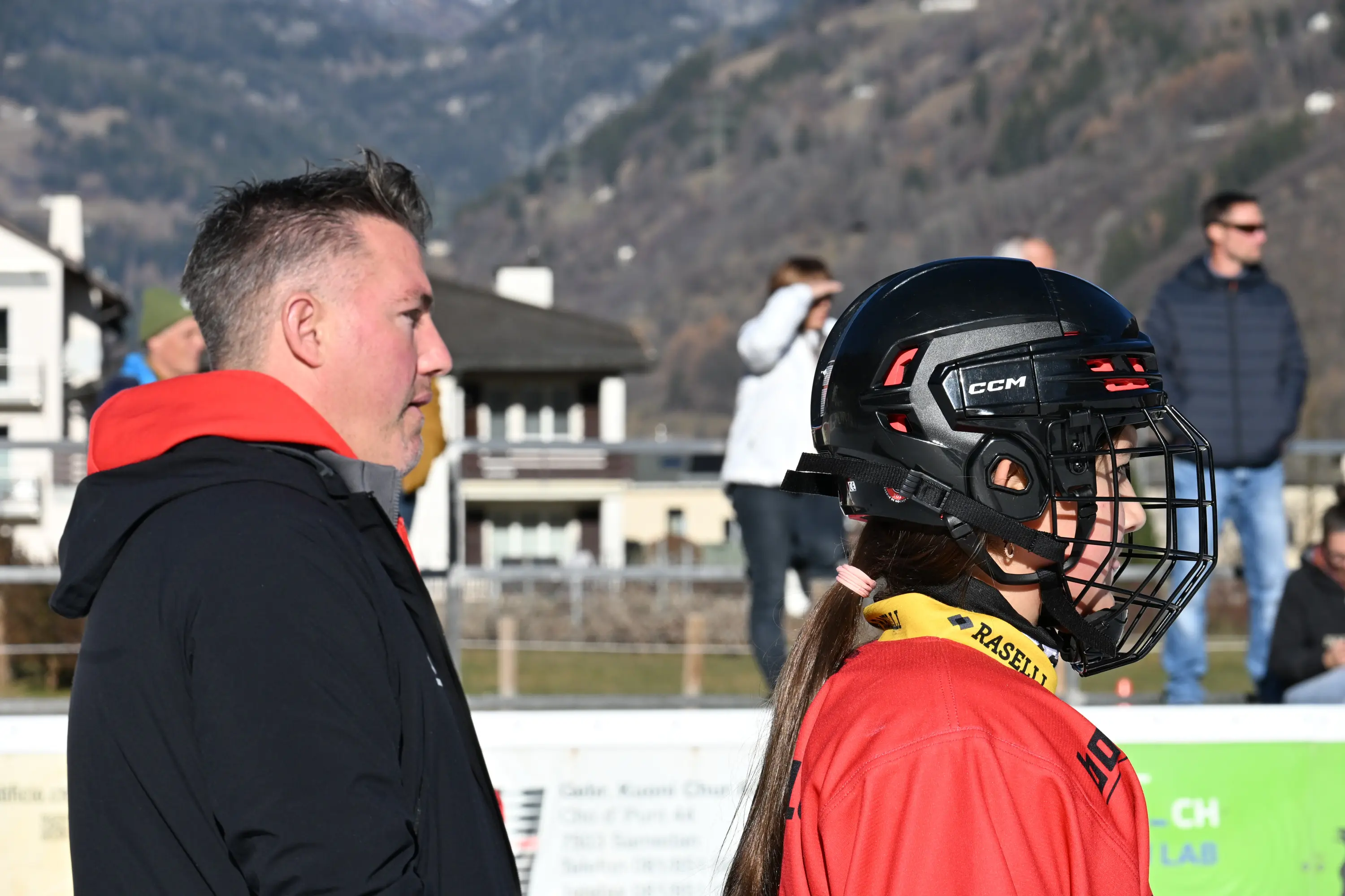Due persone in primo piano, una con un casco da hockey e l'altra con giacca nera. Sullo sfondo si vede un paesaggio montano e un gruppo di persone. È