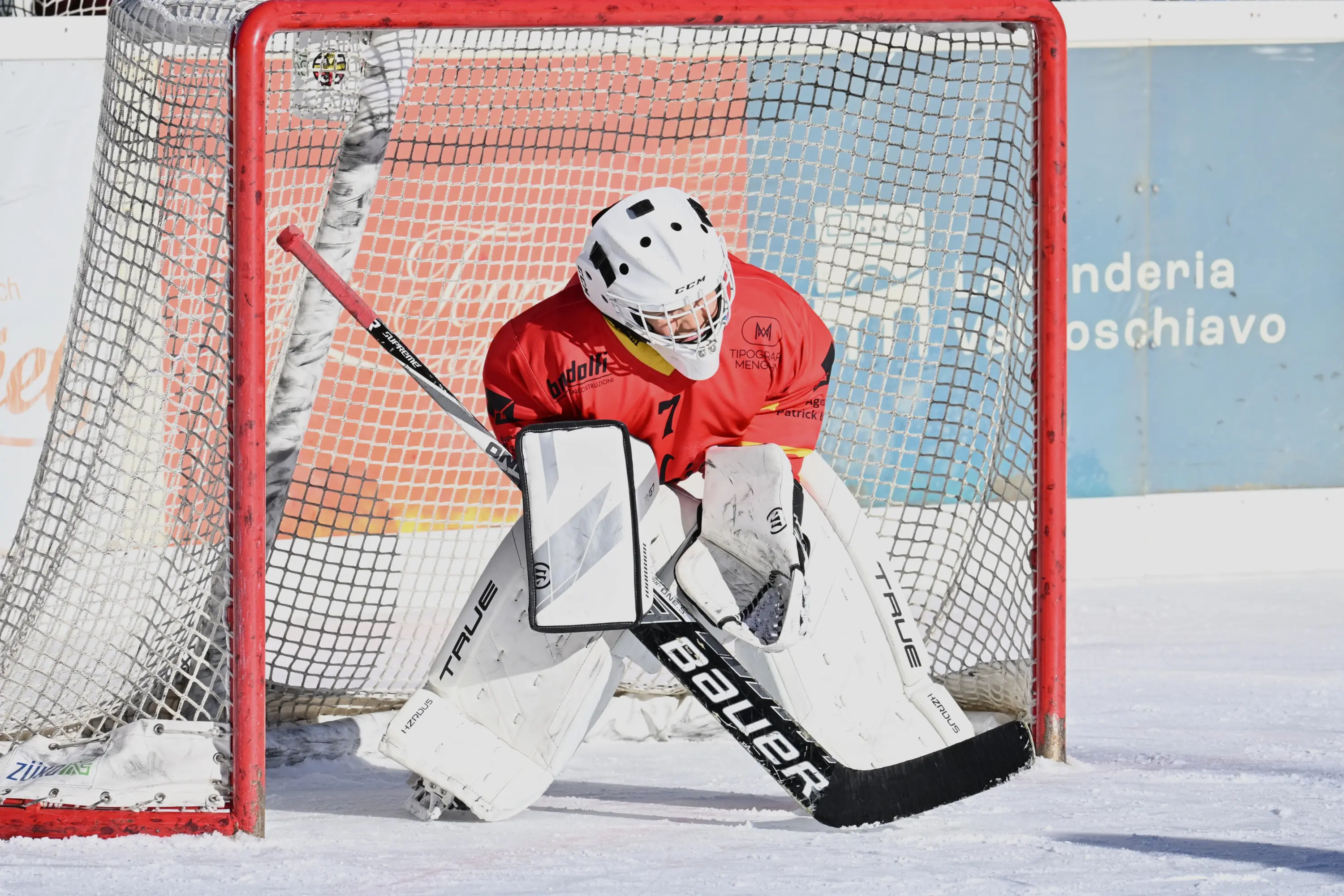 Un portiere di hockey su ghiaccio si prepara nella sua area di porta, indossando una divisa rossa e attrezzatura bianca. La rete è rossa e sul ghiaccio