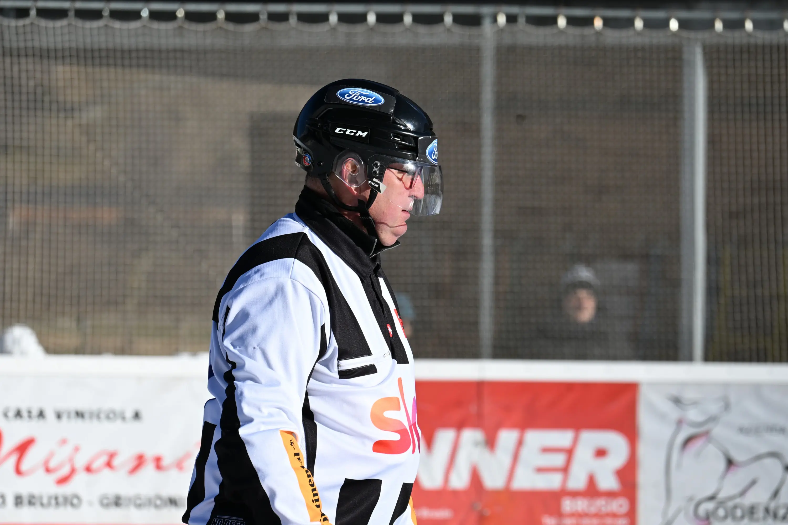Arbitro di hockey su ghiaccio in divisa, con casco e occhiali, mentre osserva il gioco. Sullo sfondo si intravede una persona e segnapunti pubblic