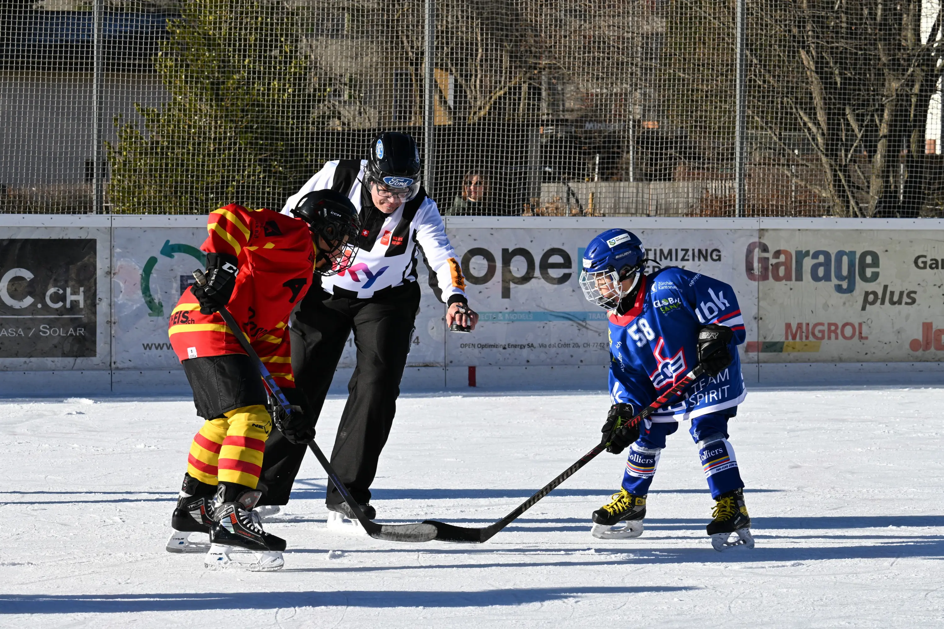 Due giovani giocatori di hockey su ghiaccio si preparano per un ingaggio, con un arbitro che osserva attentamente. Uno indossa una maglia rossa e gialla, l