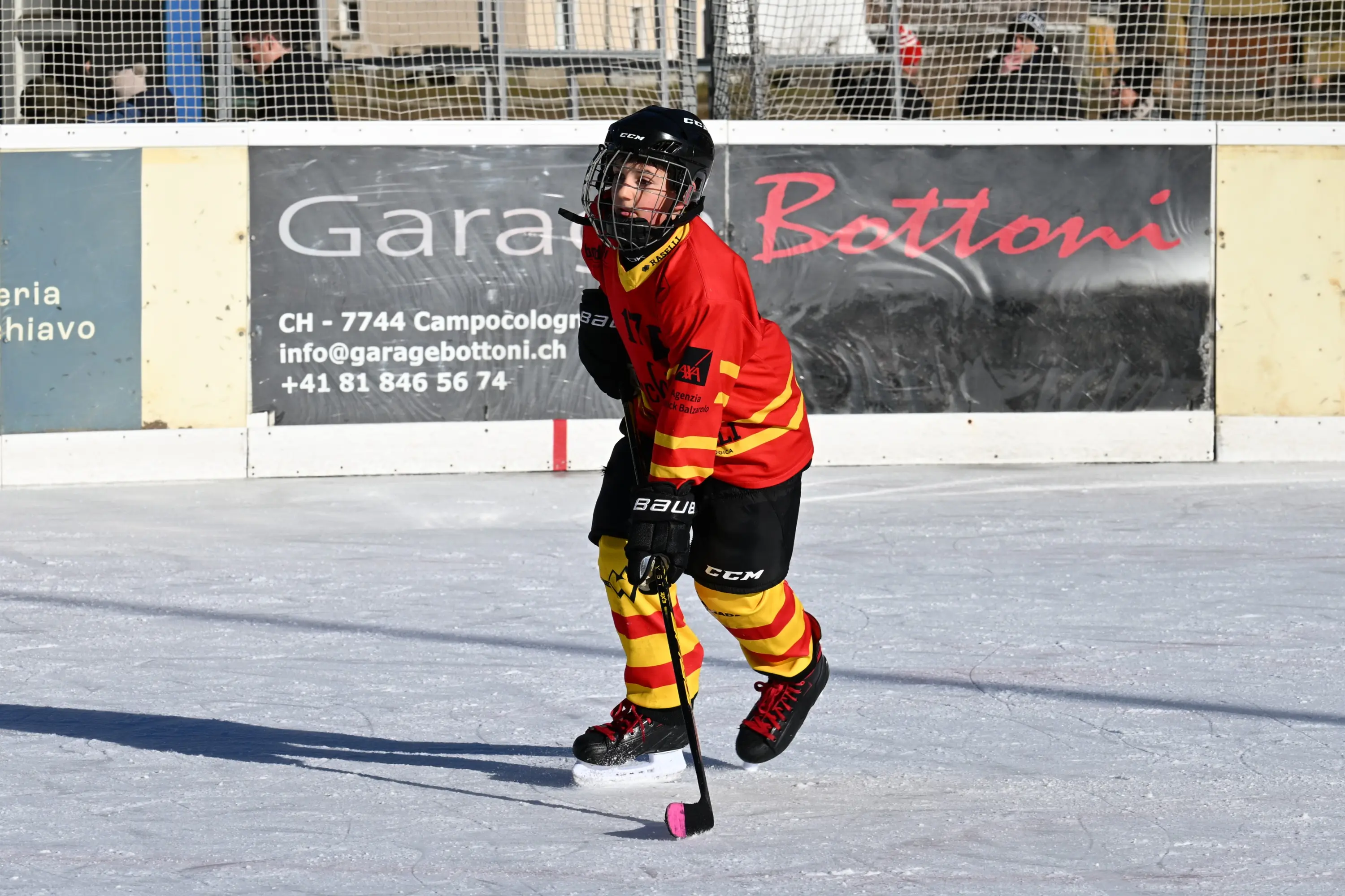 Giovane hockeista su ghiaccio, vestito con una maglia rossa e gialla, mentre pattina e tiene in mano un bastone da hockey con una pala rosa.
