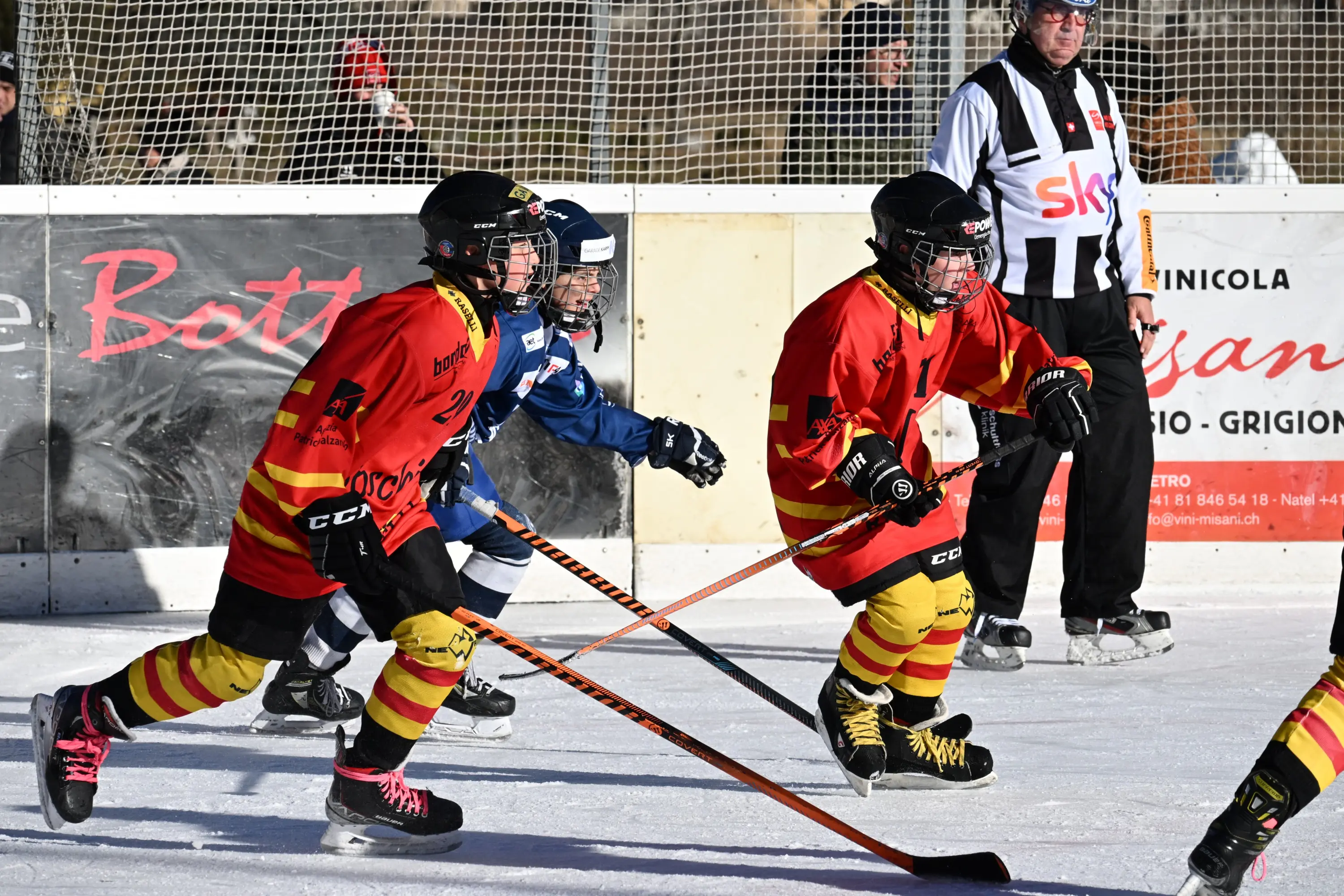Giocatori di hockey su ghiaccio in azione, indossando maglie rosse e gialle, mentre un arbitro osserva in background.