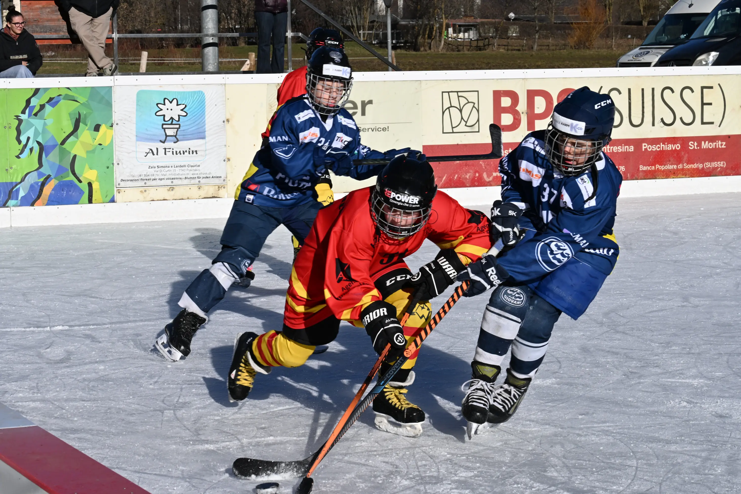 Giocatori di hockey su ghiaccio in azione su un campo all'aperto. Un giocatore con maglia rossa cerca di controllare il puck mentre viene attaccato da due av