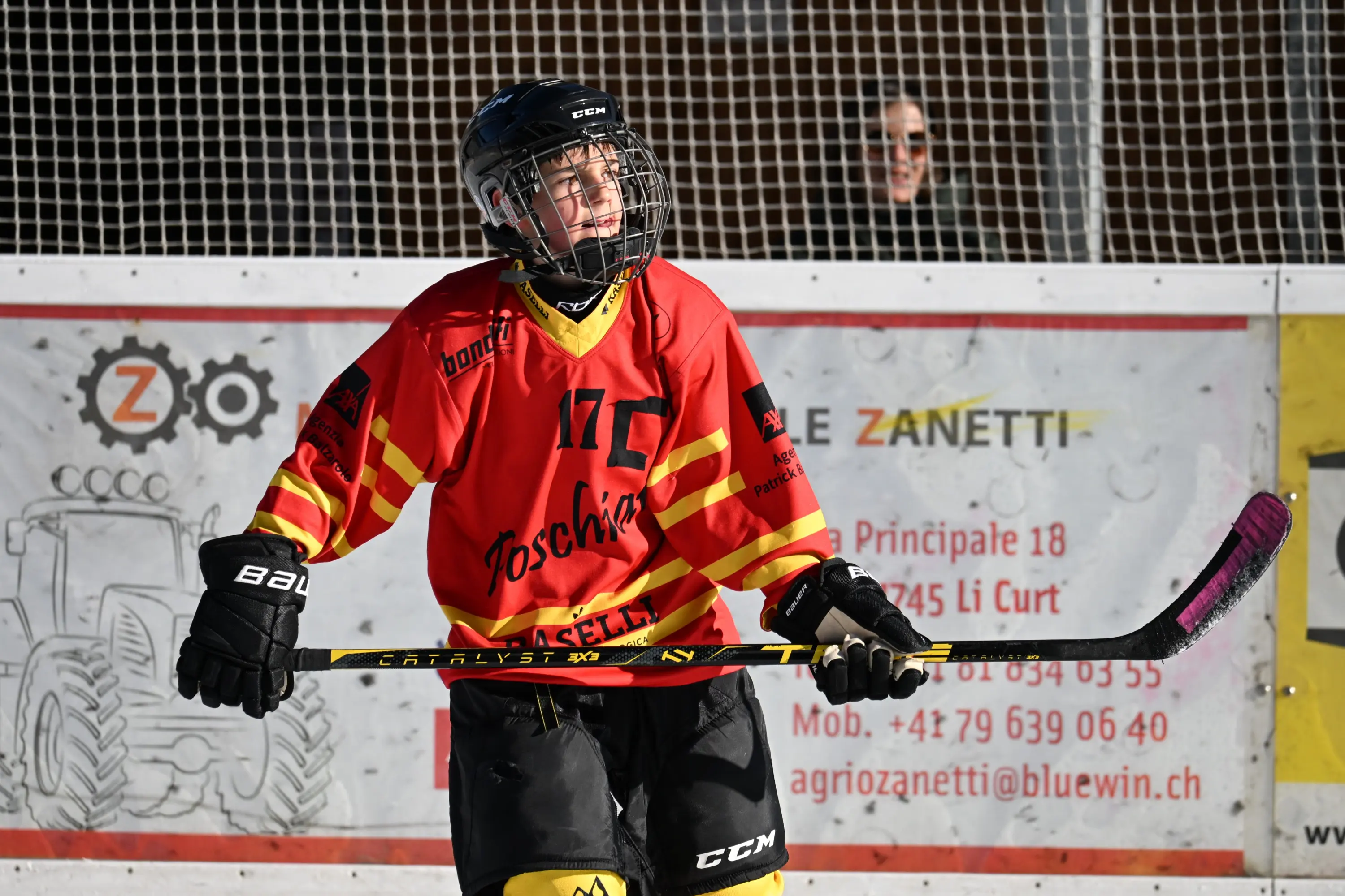 Giocatore di hockey su ghiaccio con maglia rossa e nera, indossa un casco e tiene in mano un bastone da hockey, mentre guarda verso l'alto con espression